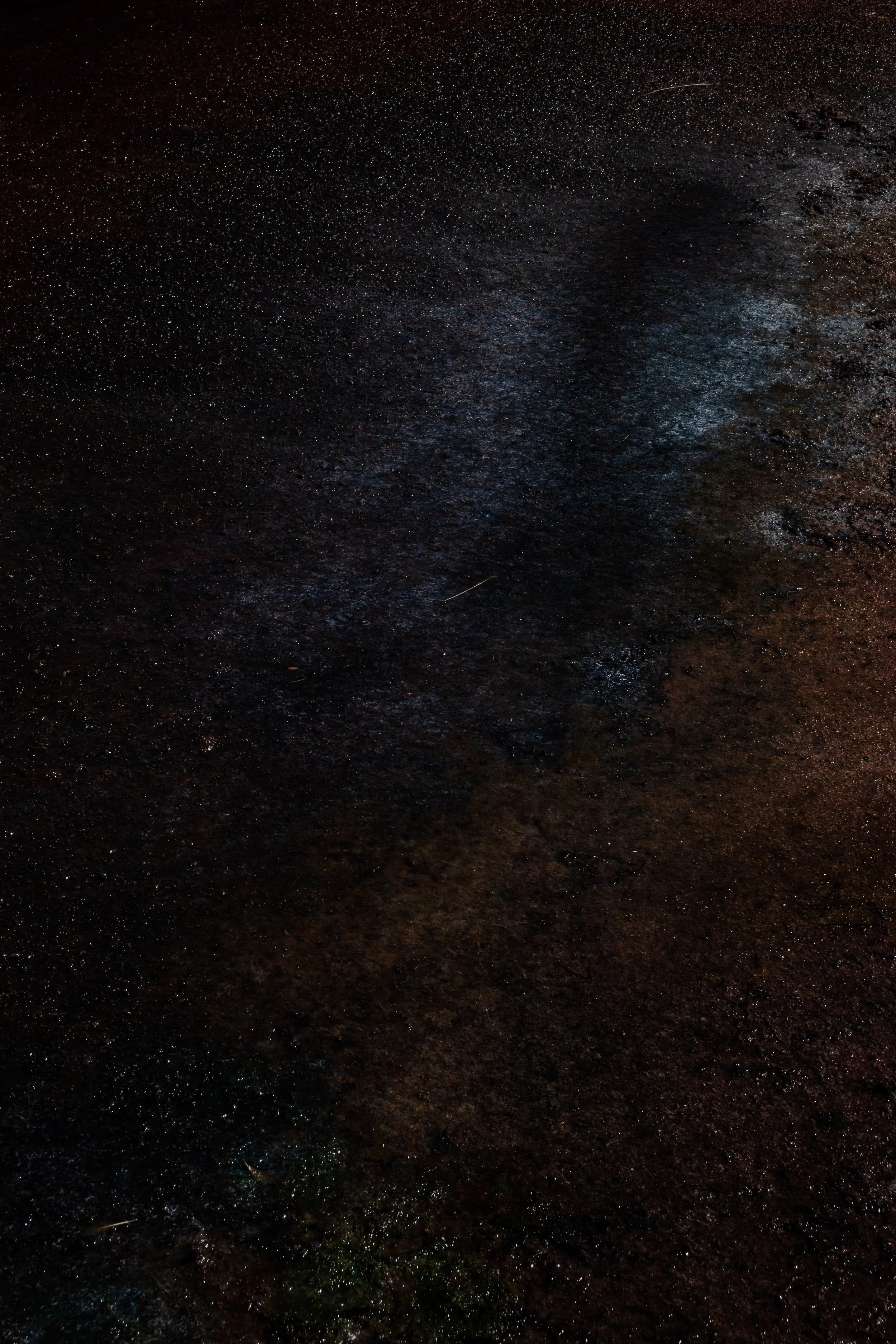Dark, textured surface with subtle blue and brown hues, possibly wet ground or pavement at night.