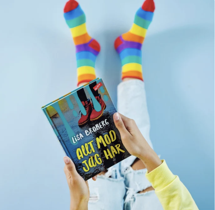 Person i färgglada regnbågsstrumpor håller upp boken ”Ärt mod jag har” av Lisa Broberg.
