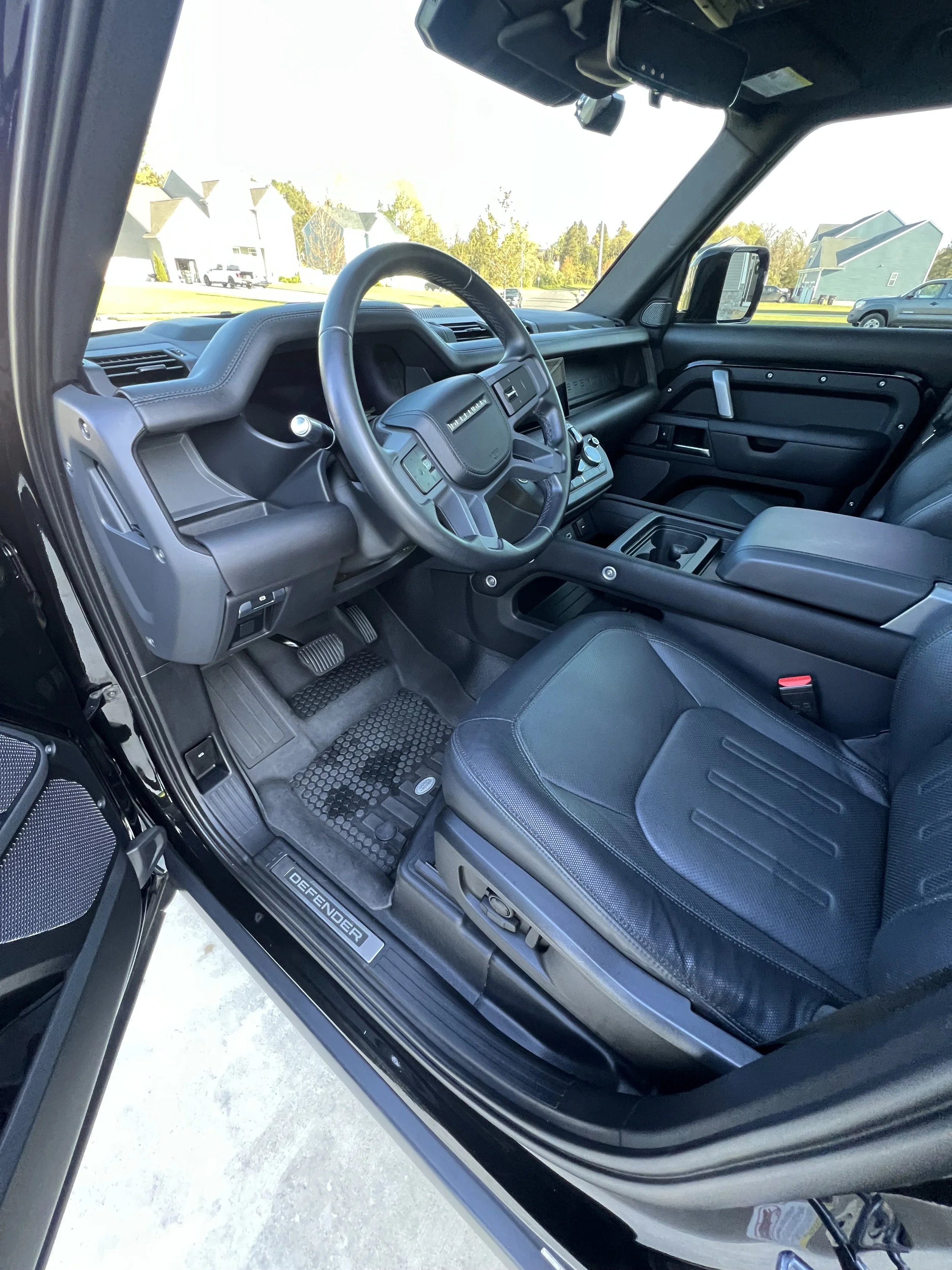 The interior of a black Ford vehicle, showing the driver's seat, steering wheel, dashboard, and console in a parking lot with buildings and trees visible outside.