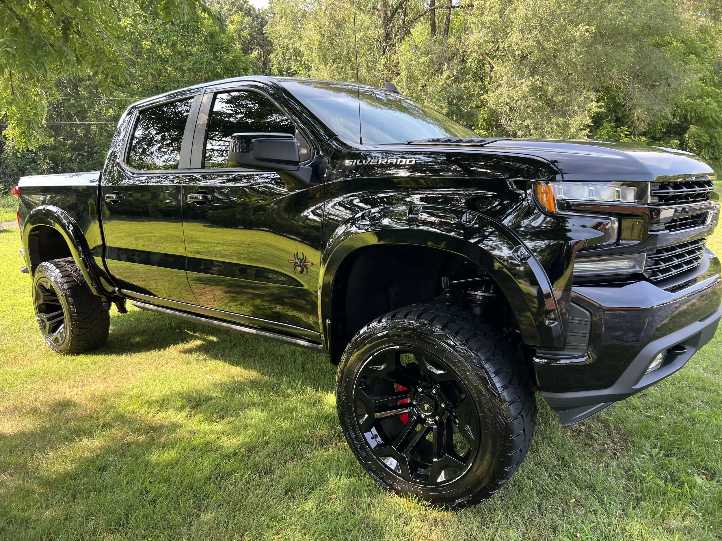 Black Chevrolet Silverado 1500 pickup truck with large off-road tires parked on grass, surrounded by trees and greenery.