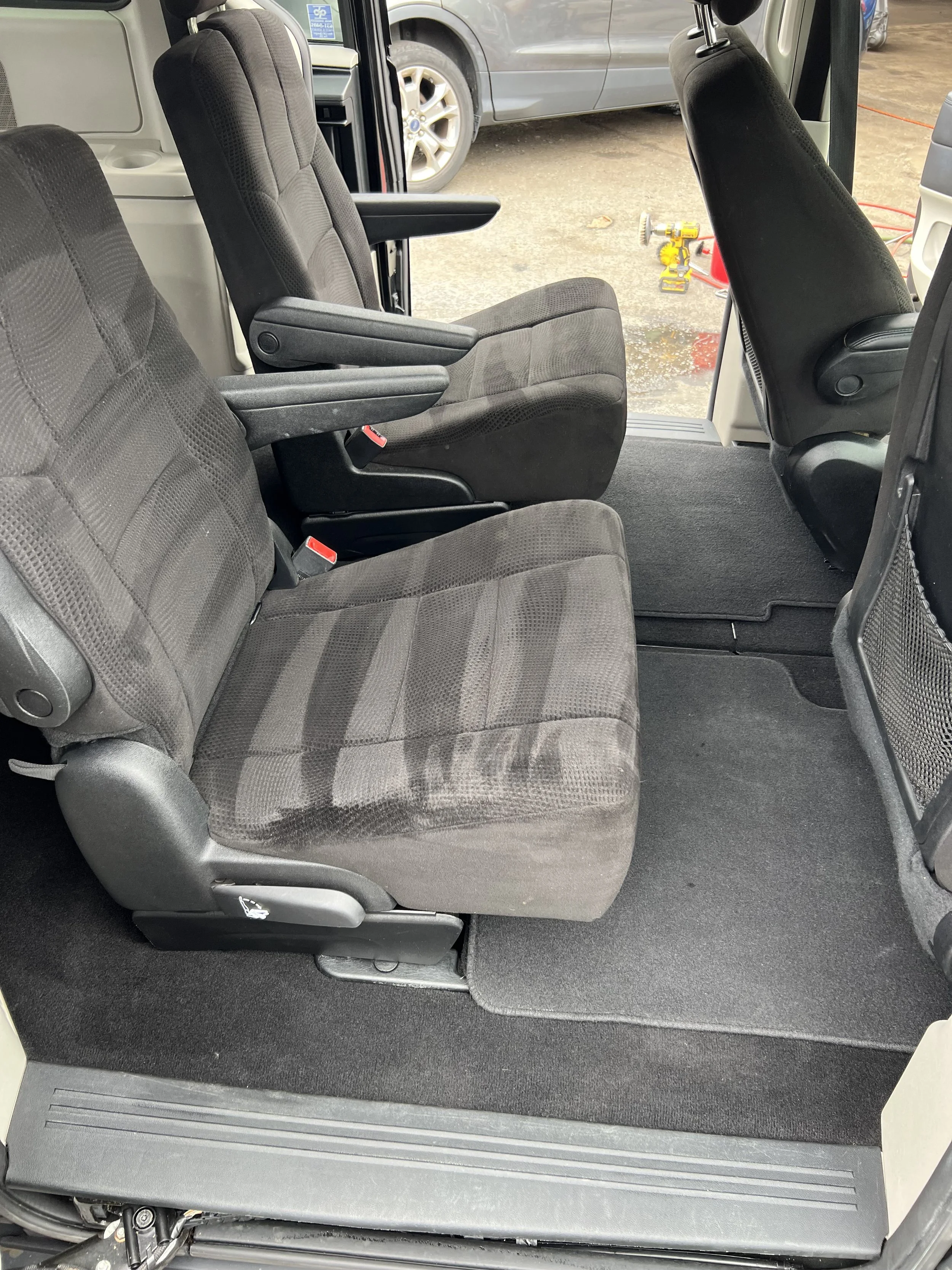 Inside of a vehicle showing two rows of empty black fabric seats with armrests, with a view outside showing parked cars and some tools.