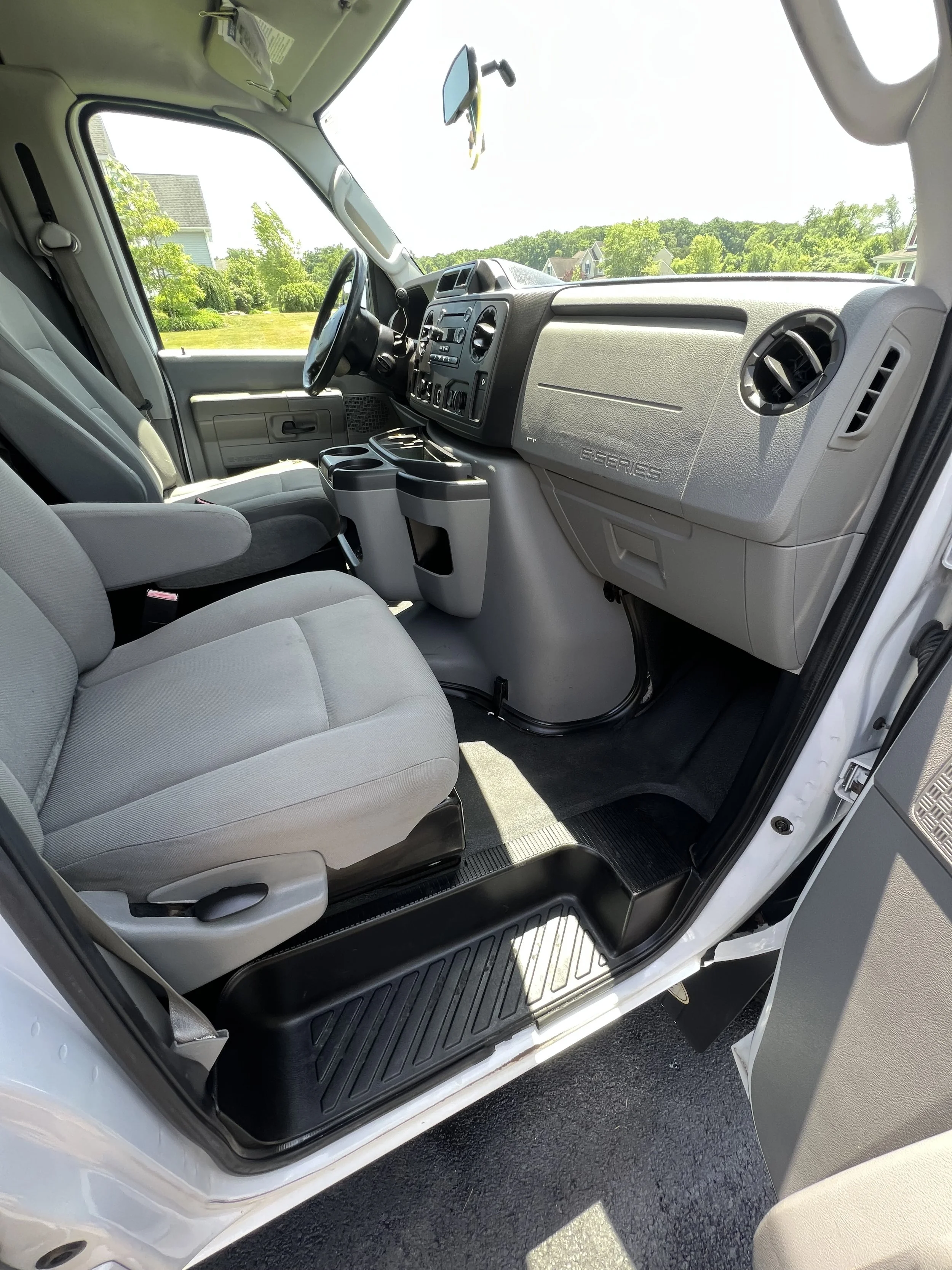 The interior front passenger and driver seats of a cargo van, with gray fabric seats, black floor mat, and dashboard with various controls, parked outdoors during daytime.