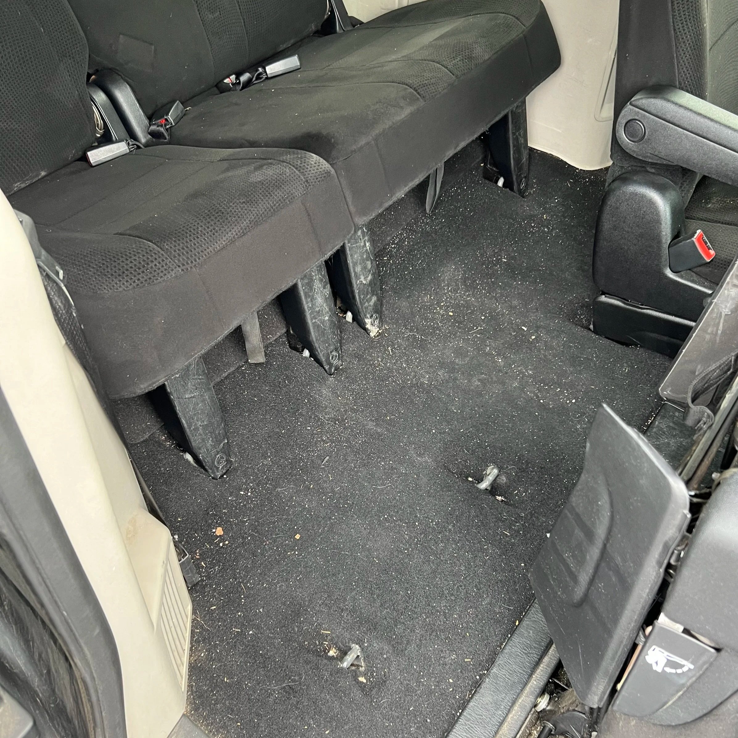 The backseat area of a vehicle with the third-row seats folded down, showing the dirty and dusty black carpeted floor.