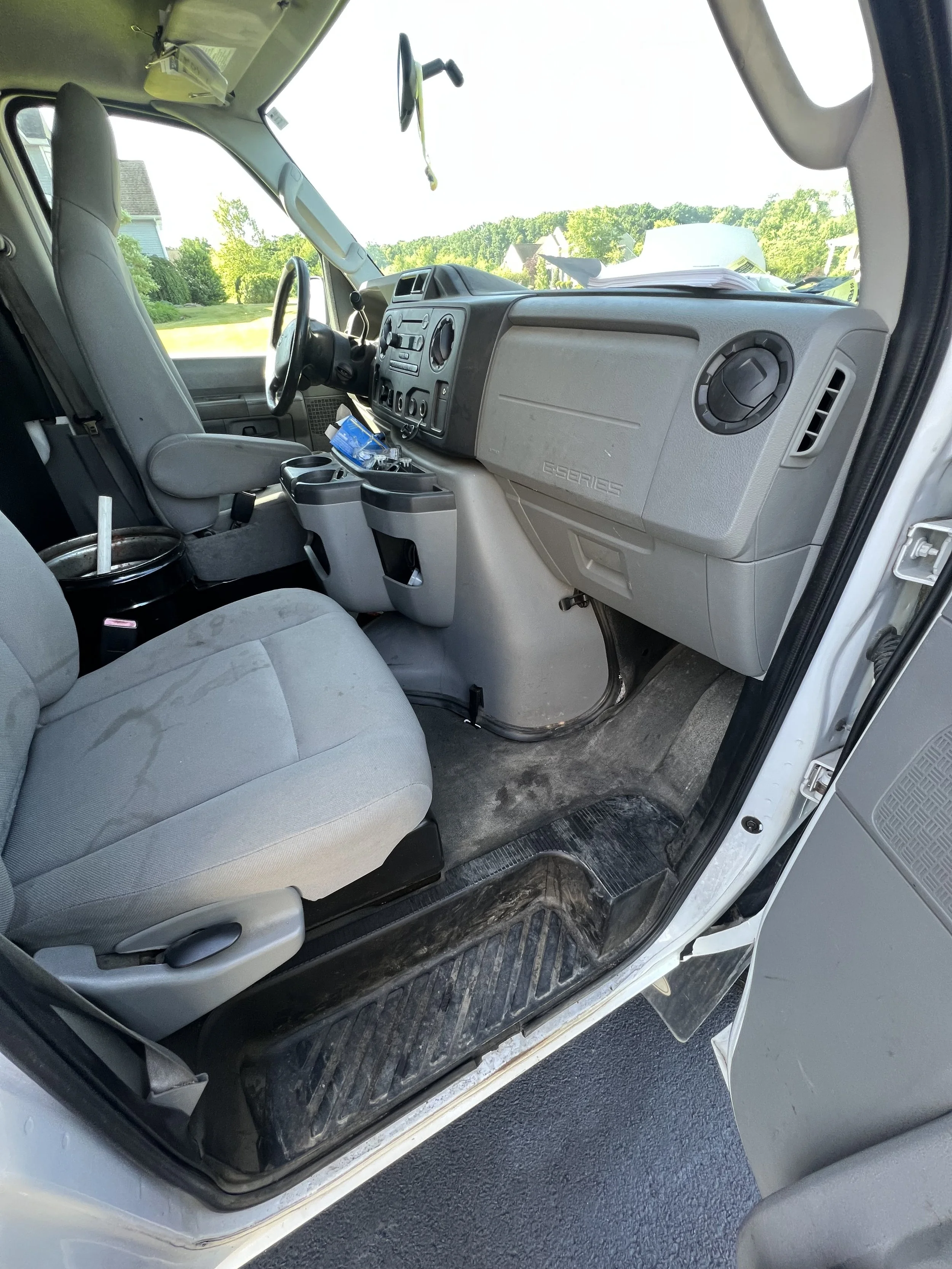 The interior of a commercial vehicle cab showing the driver's seat, dashboard, steering wheel, and various controls. The driver's seat has a cup holder with a drink, and the floor has a rubber mat. Outside the cab, a green landscape with trees and ho