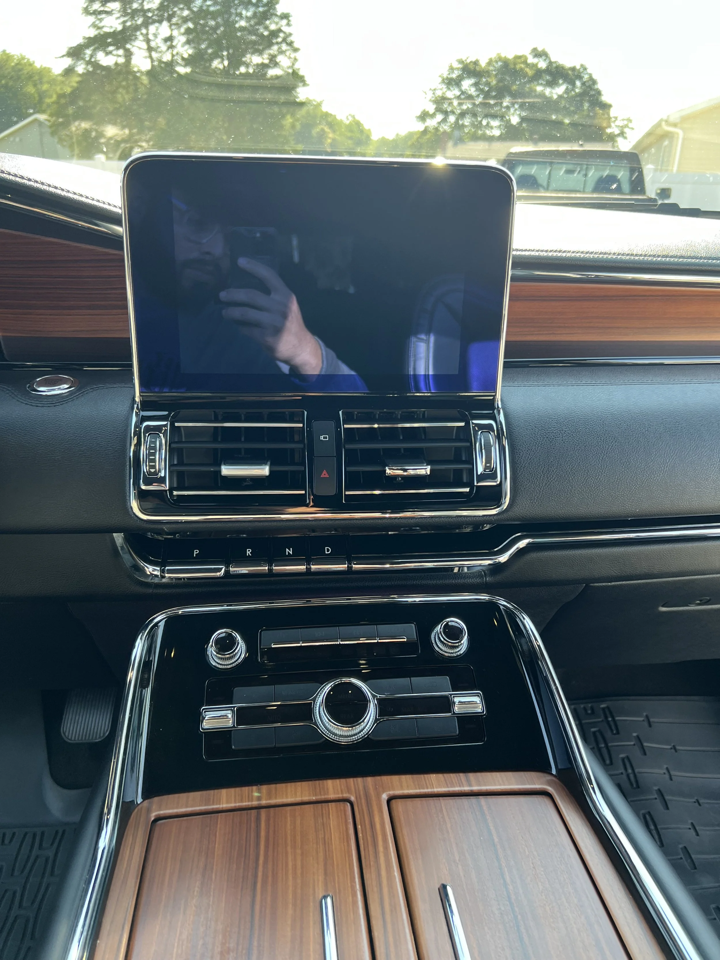 Inside view of a luxury car dashboard with a large central touchscreen, air vents, control buttons, and wood trim, with sunlight coming through the windshield.