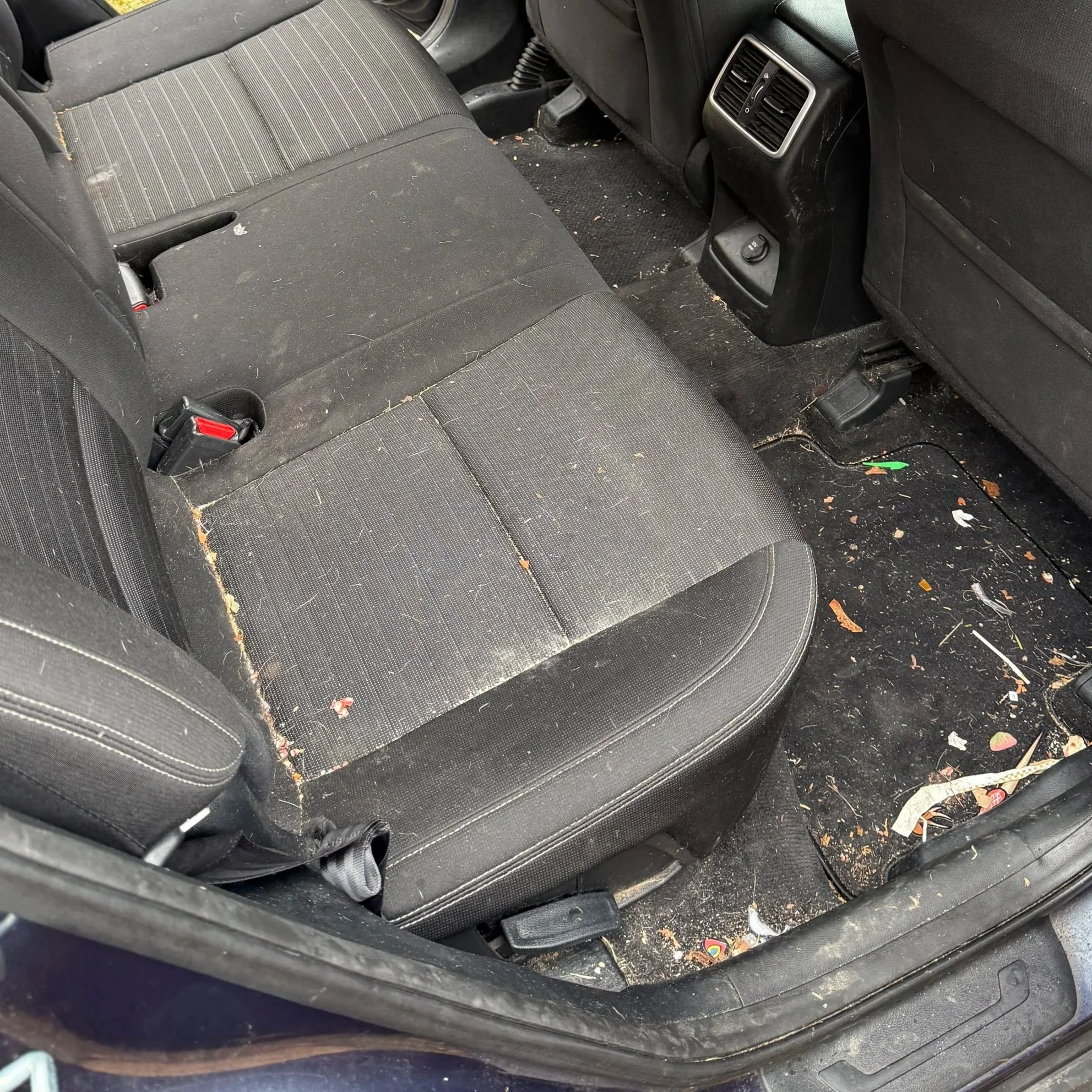 Dirty car interior with black fabric seats and floor mats, scattered with trash, crumbs, and debris.