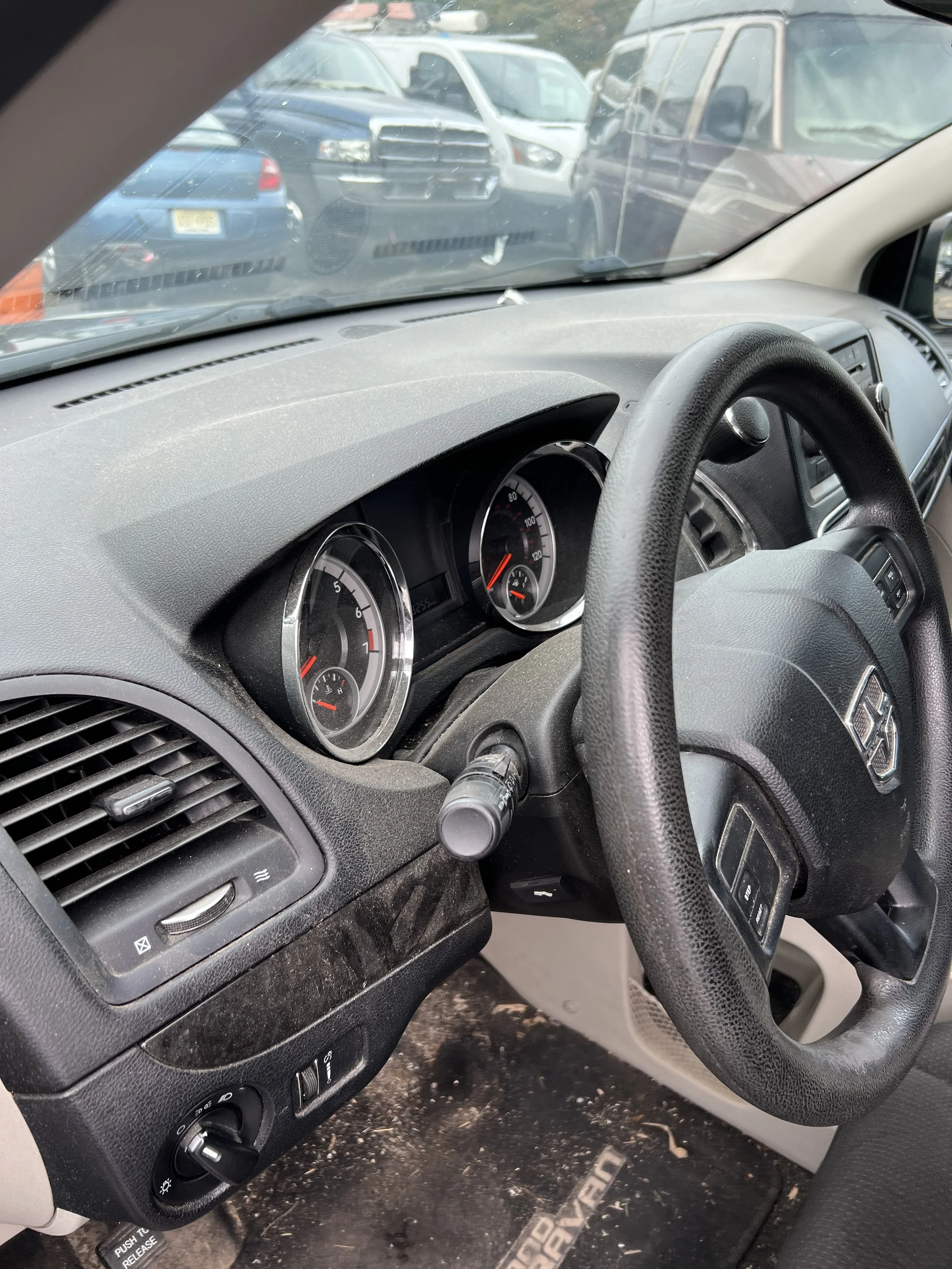 Inside of a vehicle showing the dashboard, steering wheel, and a view through the windshield of a parking lot with multiple parked cars.