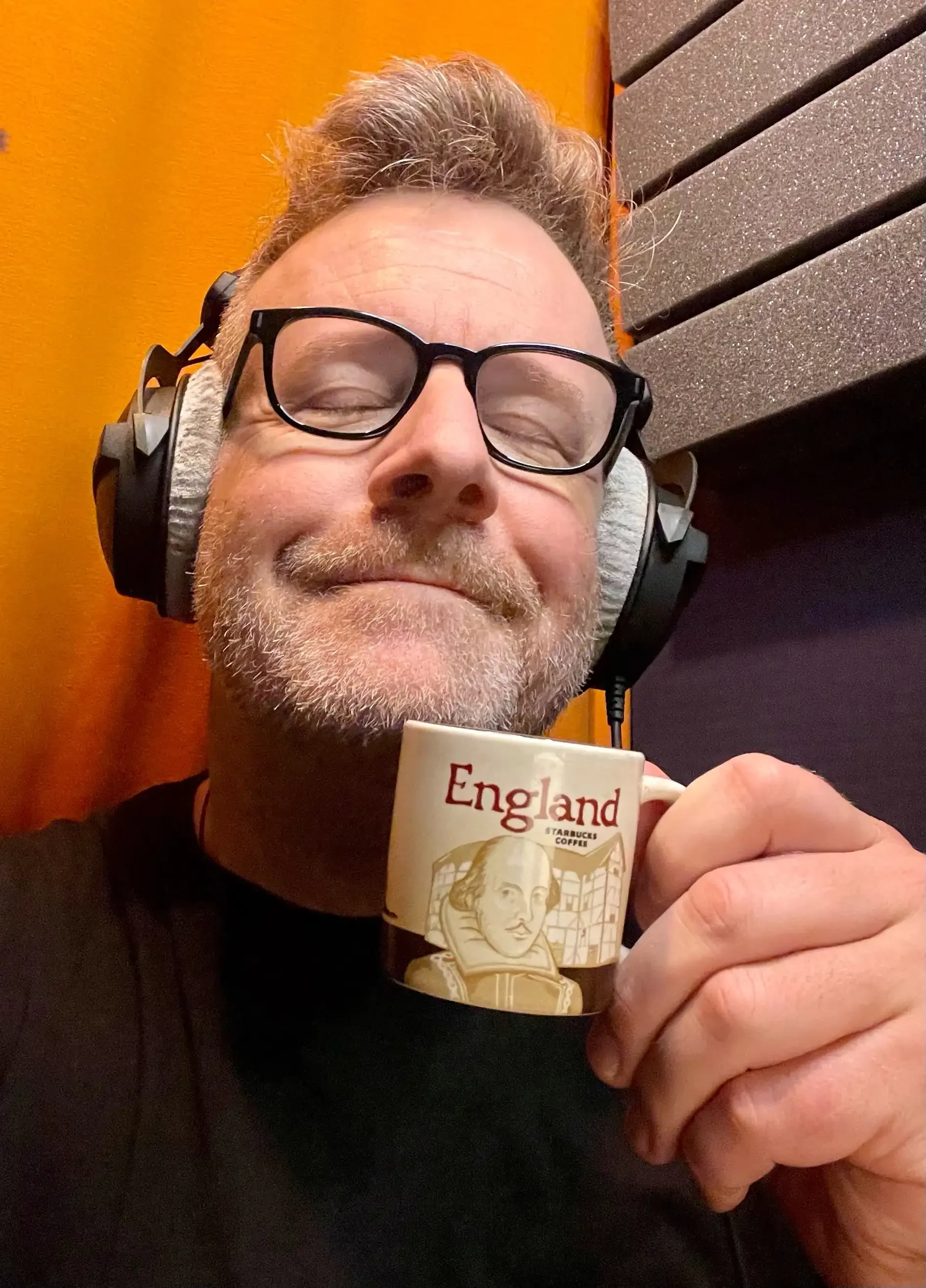 A man with glasses and a beard smiling with eyes closed, holding a coffee mug labeled 'England' with a portrait of William Shakespeare, in a recording studio with orange and black soundproofing panels.