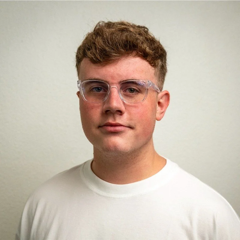 A young man with short, curly brown hair, wearing clear glasses and a plain white T-shirt, standing against a light-colored wall.