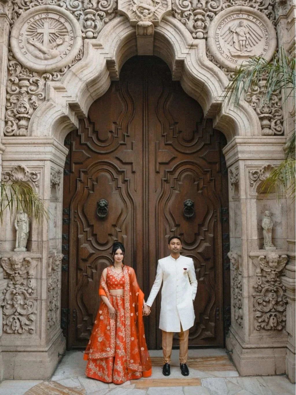 Bride and bridesmaids standing in front of a white wall with colorful tiles, holding bouquets of white flowers, dressed in pink and sparkly dresses, smiling at a wedding celebration.