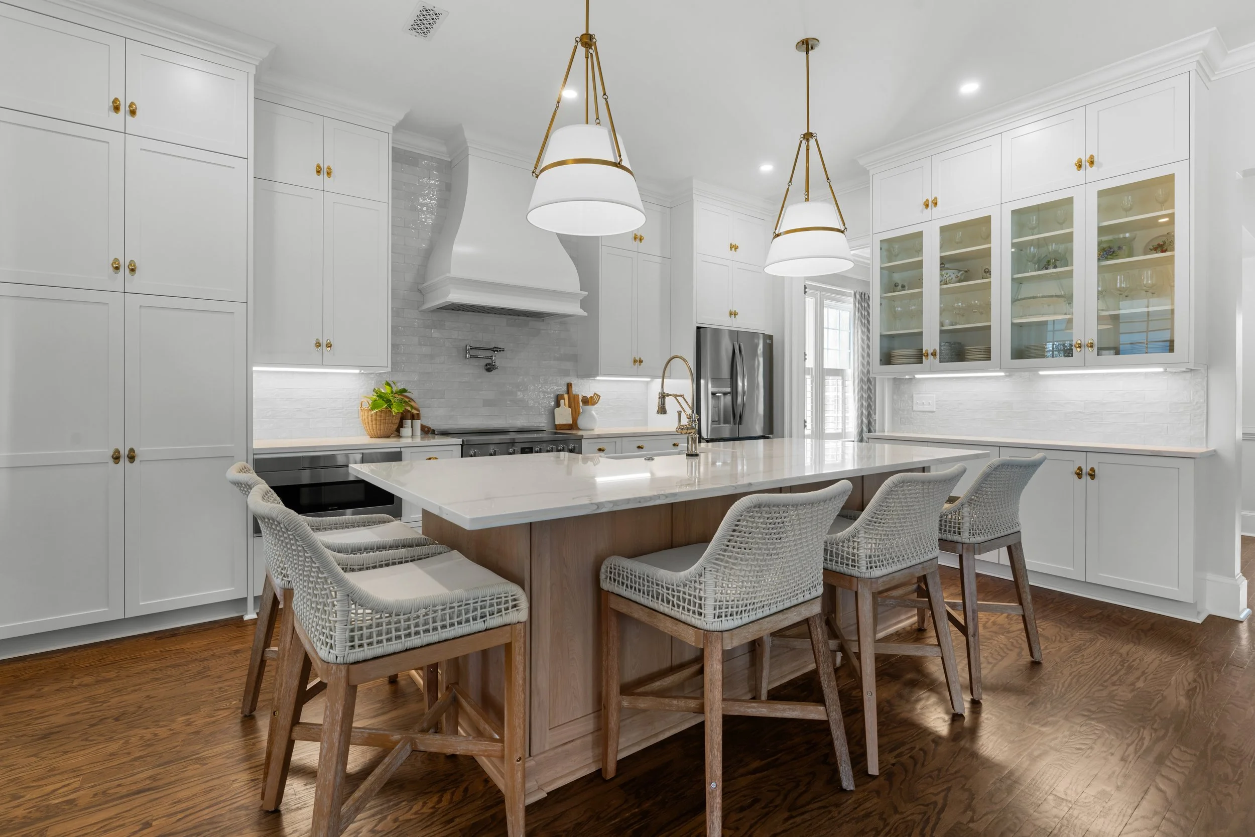 Bright modern kitchen with white cabinets, a large center island with a marble countertop, four barstools with wicker seats, pendant lights, stainless steel appliances, and hardwood floors.