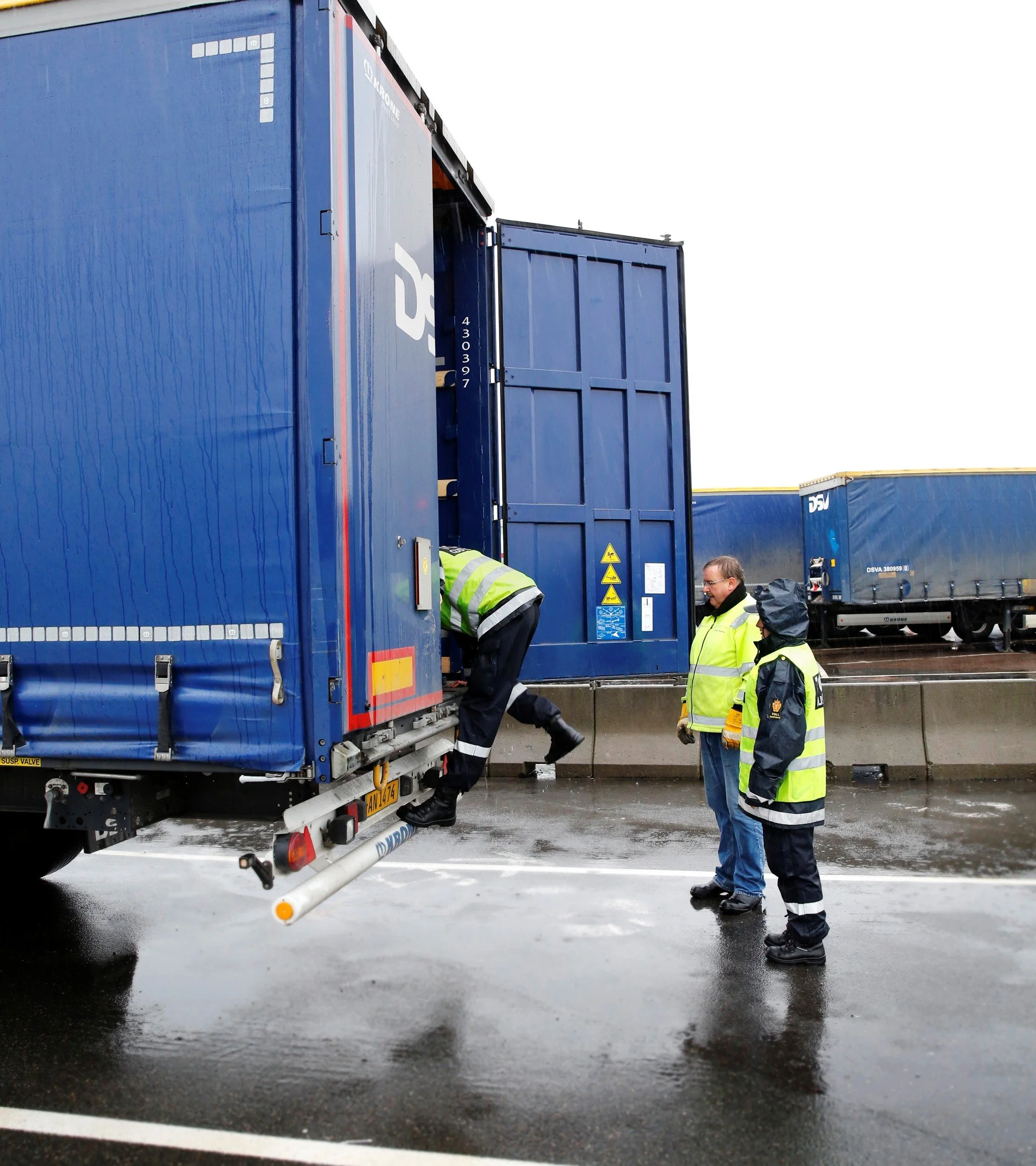Personer i refleksvester jobber med å koble til en stor blå lastebil i dårlig vær, med våt asfalt og regn.
