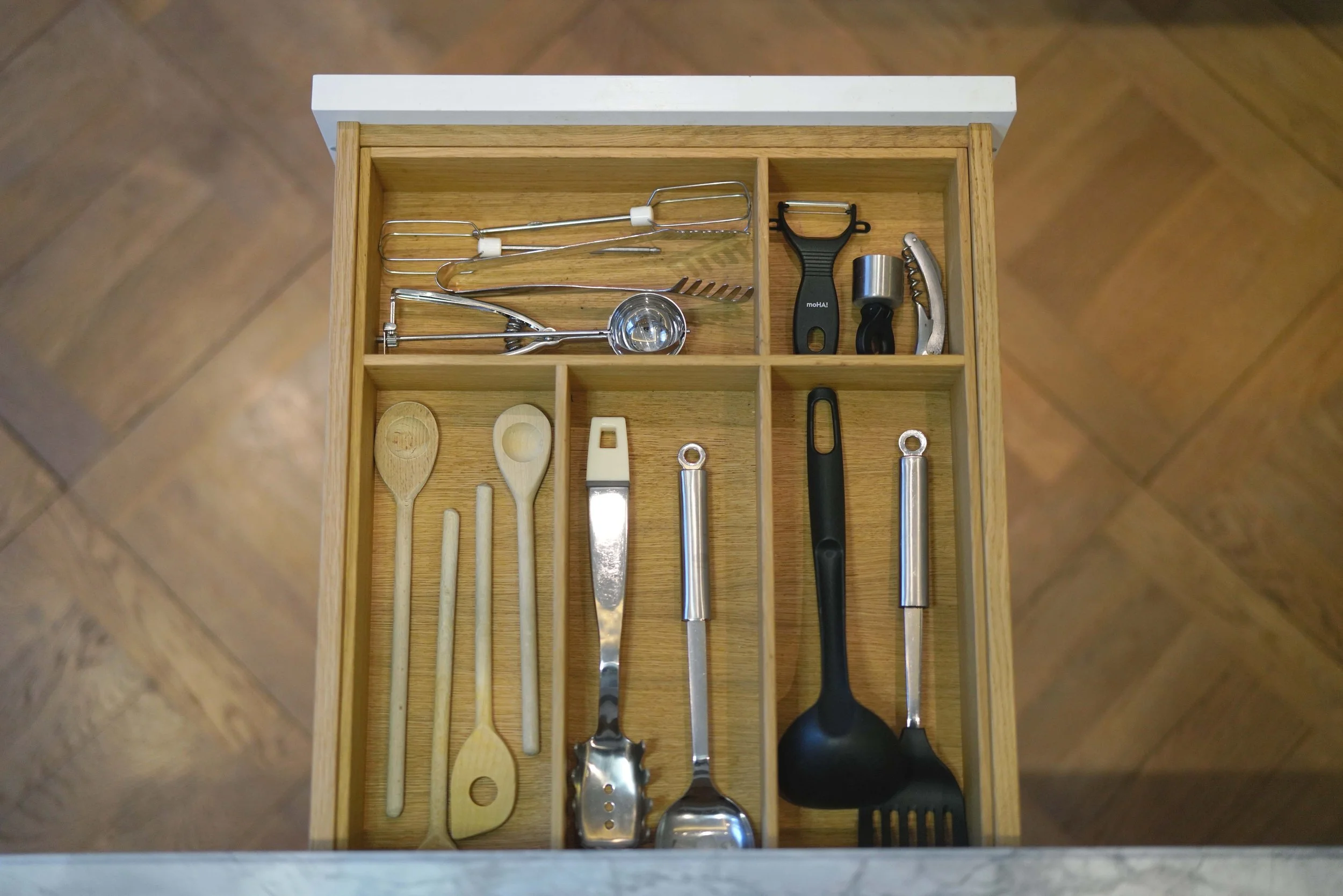 Küchenutensilien in einer kleinen Holzschublade, darunter Holzlöffel, Schneebesen, Korkenzieher, Messer, Pfannenwender und andere Kochwerkzeuge.