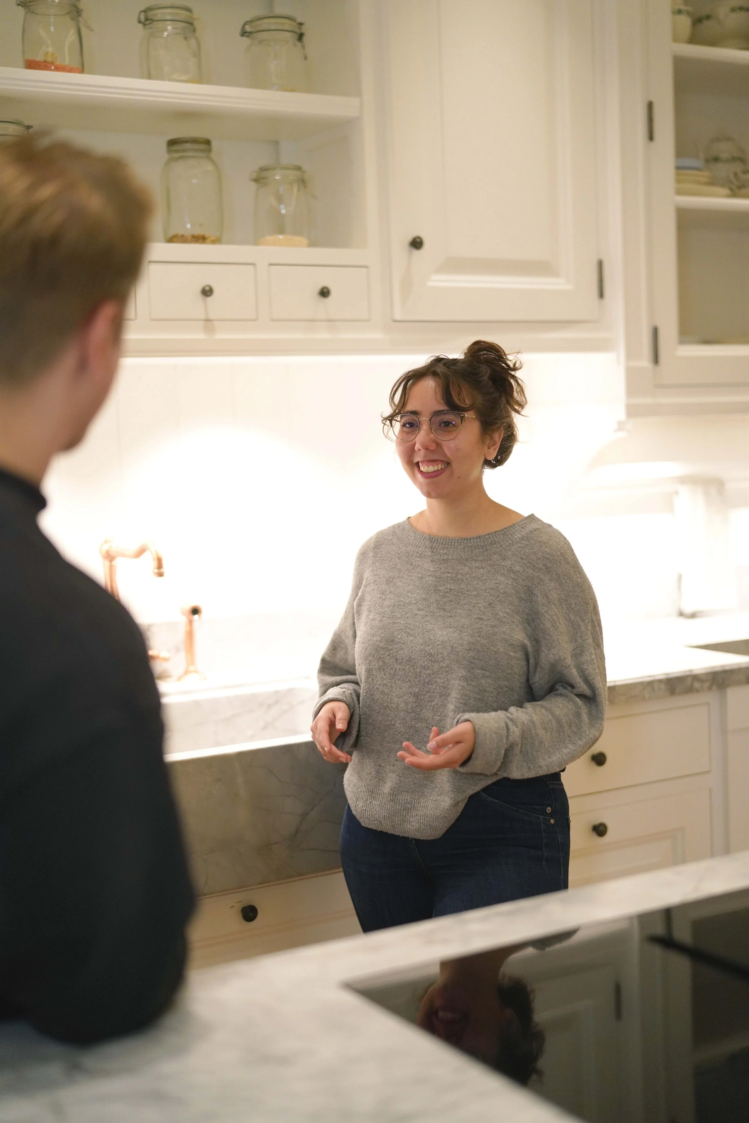 Eine lachende Frau mit Brille und grauem Pullover spricht mit einem Mann in dunkler Kleidung in einer weißen Küche.