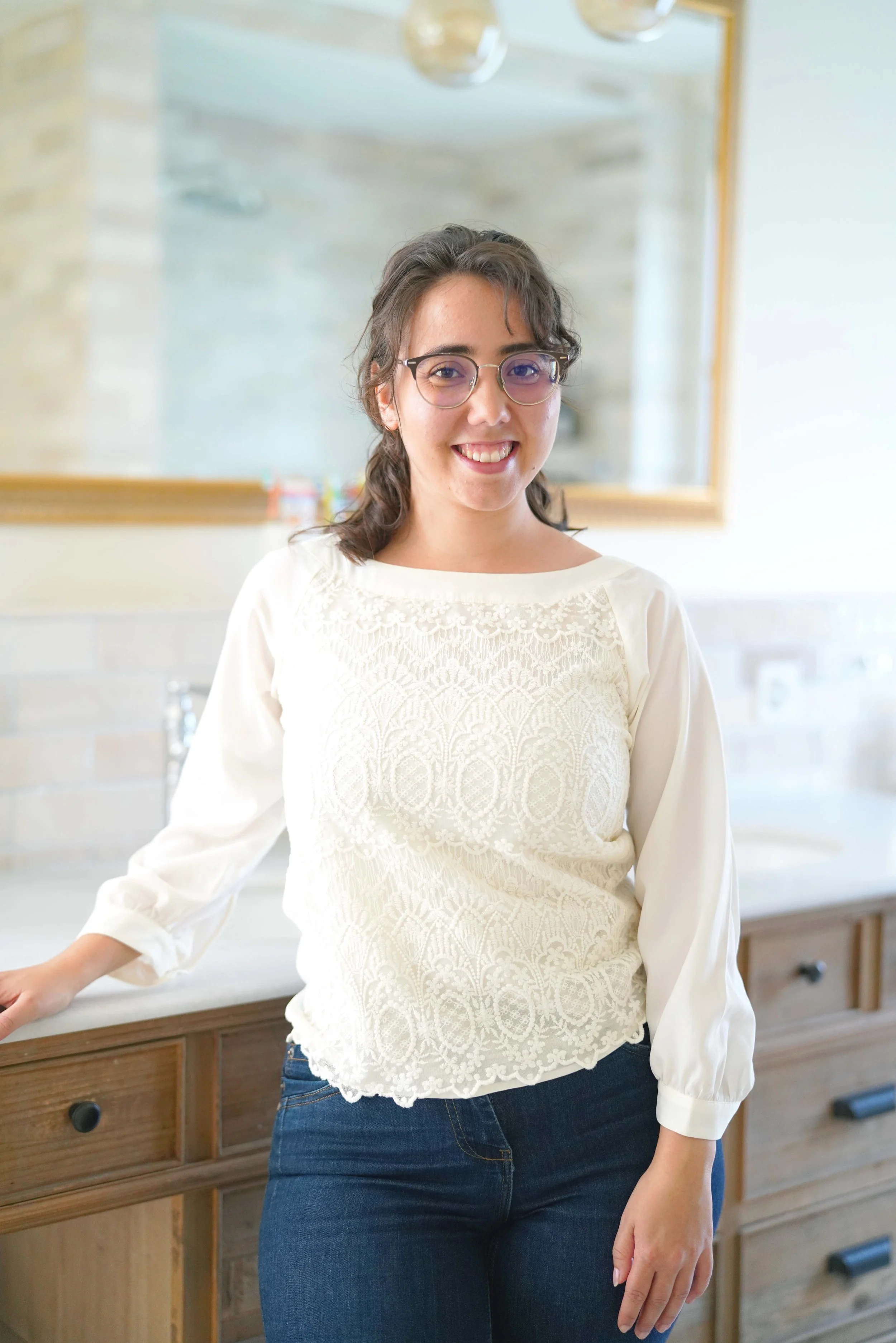 Junge Frau mit Brille und lockigem Haar, lächelnd, steht in einem modernen Badezimmer.