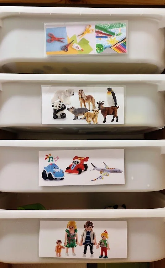 Vier weiße Schubladen mit Fotos. Das obere Foto zeigt Bastelmaterialien, das zweite Foto Spielzeugtiere, das dritte Foto Spielzeugautos und Flugzeuge, das vierte Foto Puppen und ein Kinderspielzeug, das unterste Foto zeigt zwei Puppen und zwei Kinderfiguren.