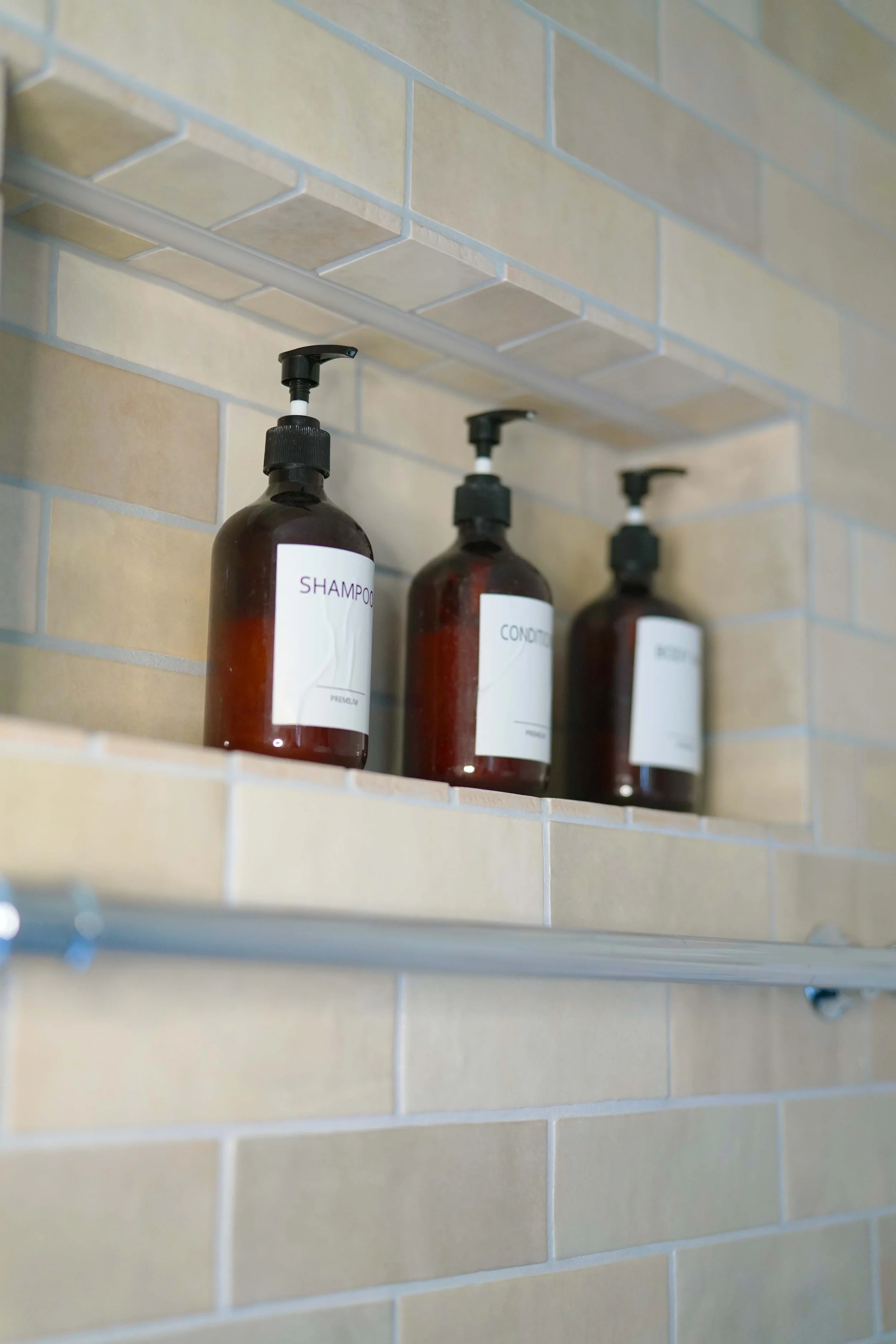 Drei braune Shampoo- und Conditioner-Flaschen mit Pumpenspendern in einer Nische in einer Badezimmerwand aus beige Fliesen.