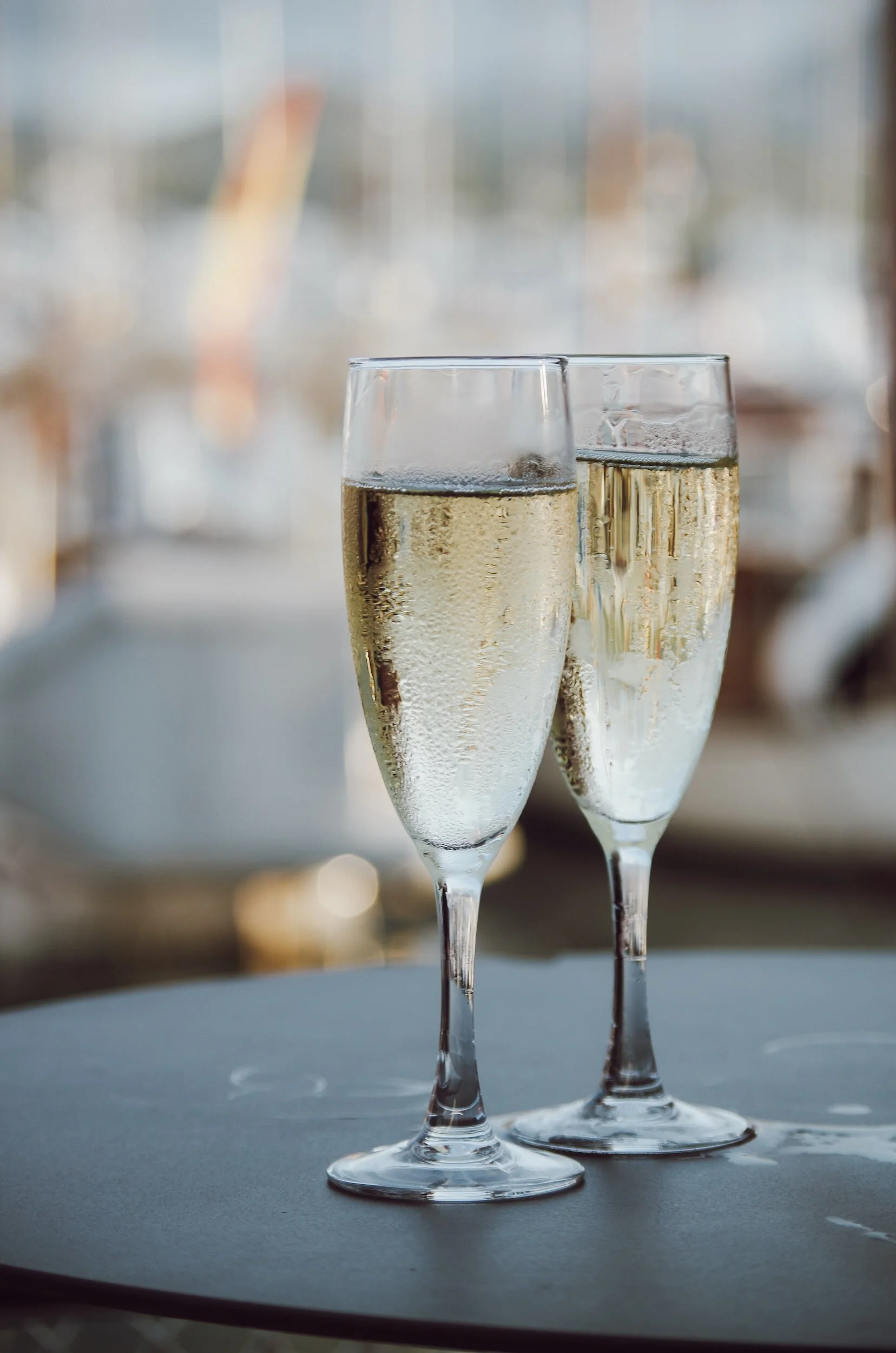 Two champagne flutes filled with sparkling white wine on a table with a blurred outdoor background.