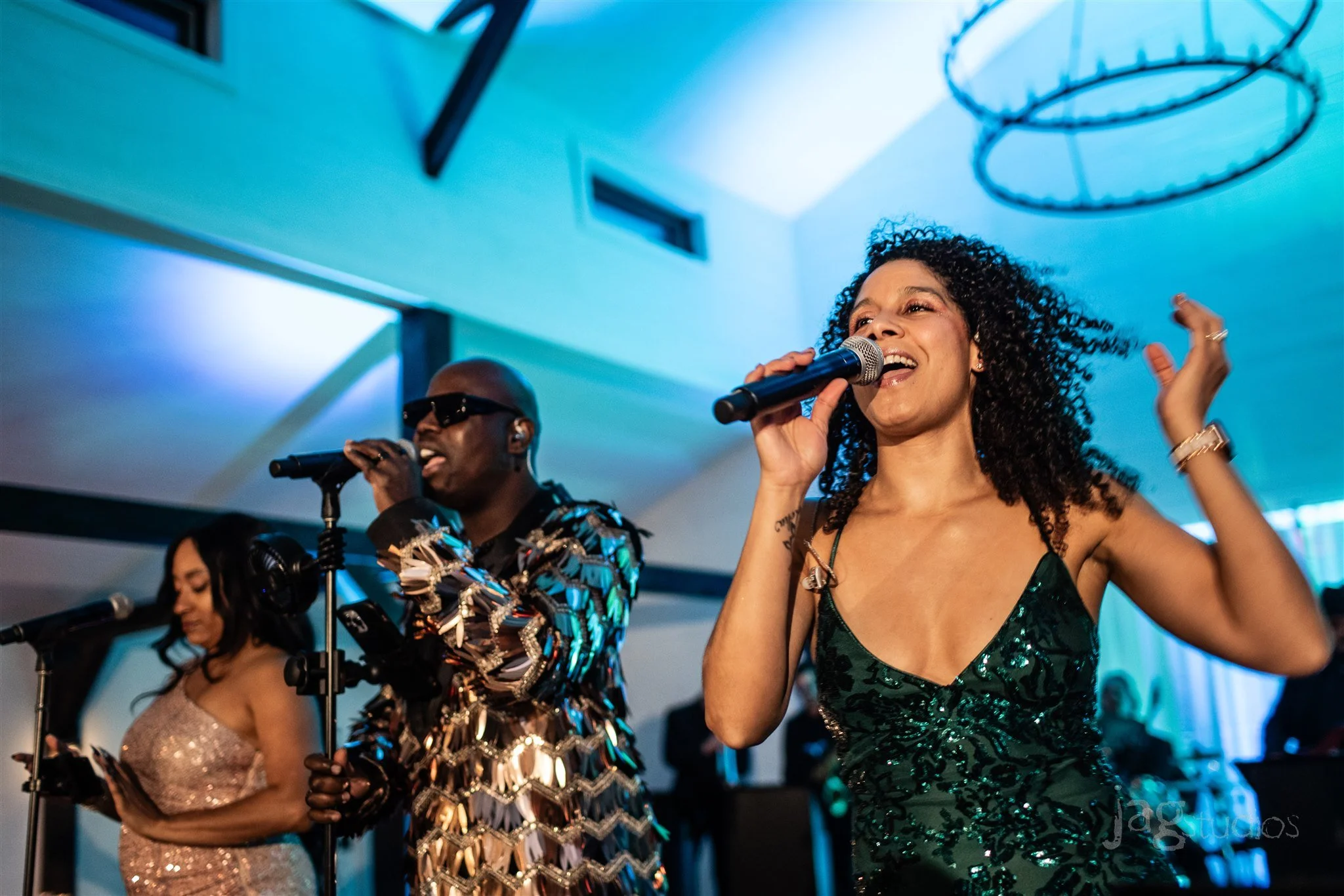 Three musicians performing on stage, each holding a microphone, with colorful lighting and a modern ceiling in the background while Eido Bar Company bartends the after party.