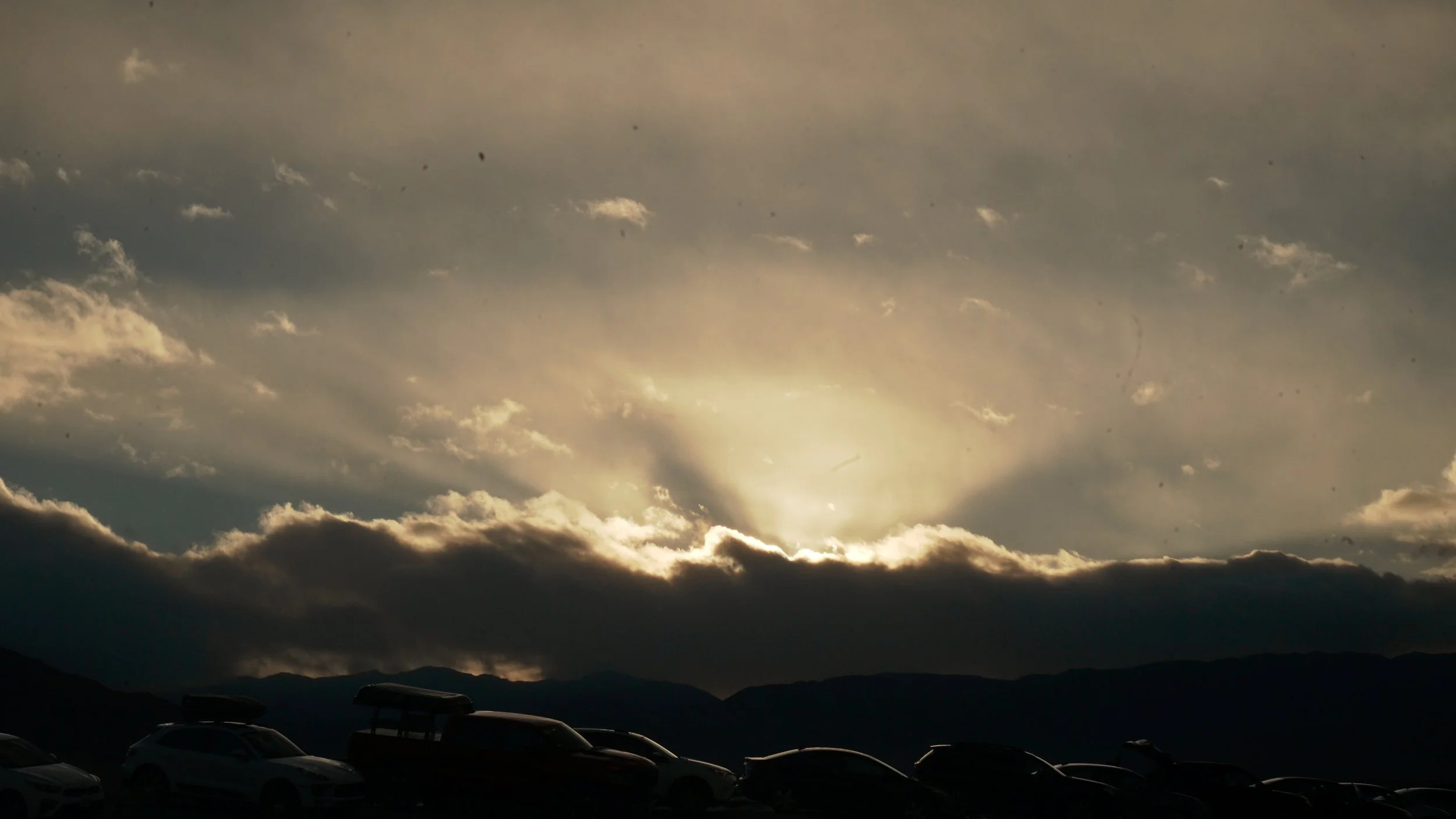Silhouette of cars parked in front of a mountain range during sunset with rays of sunlight shining through clouds.