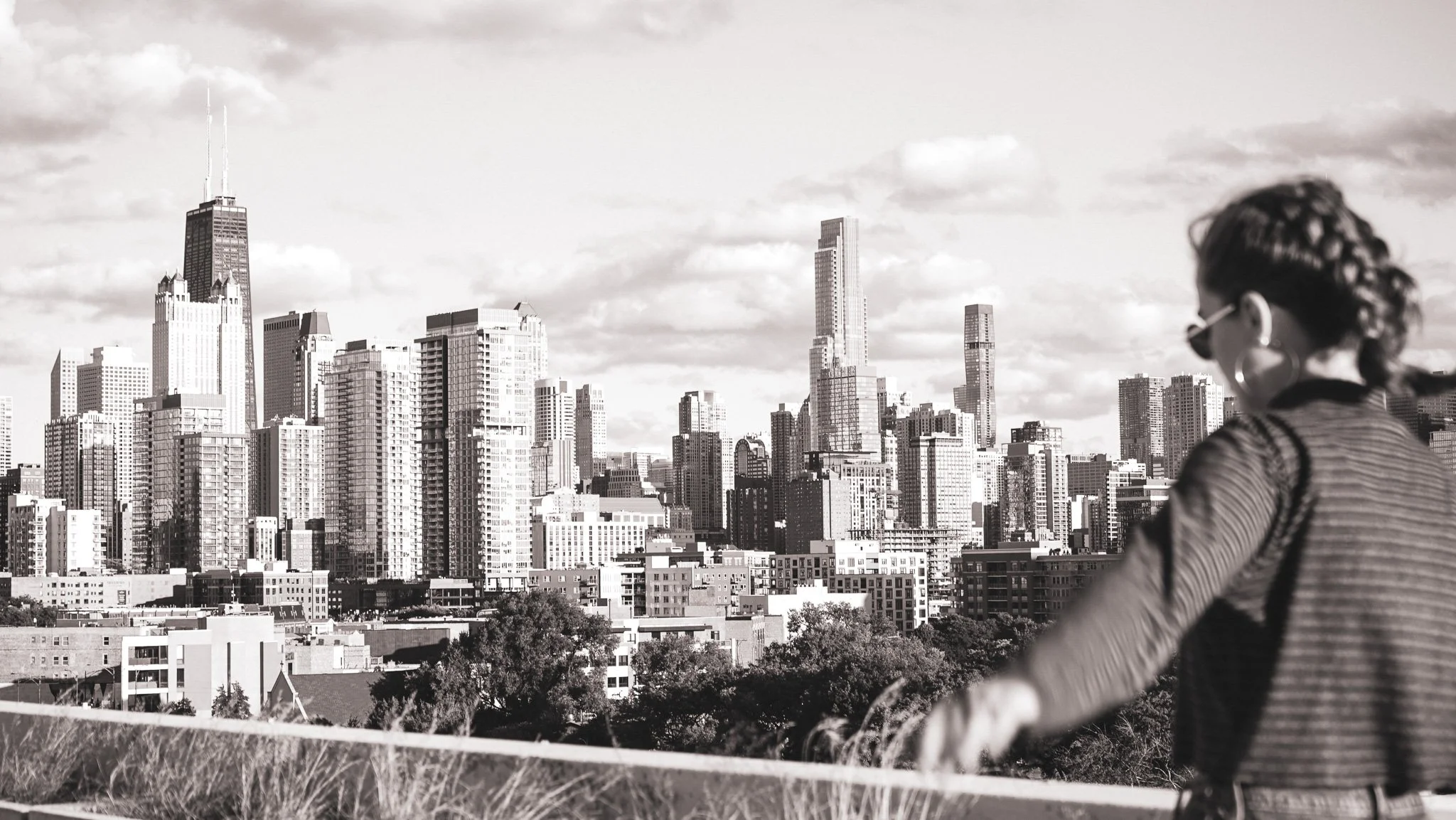 A woman wearing sunglasses and a striped jacket is standing in front of the Chicago skyline, with tall buildings and skyscrapers visible in the background, in a black and white photograph.