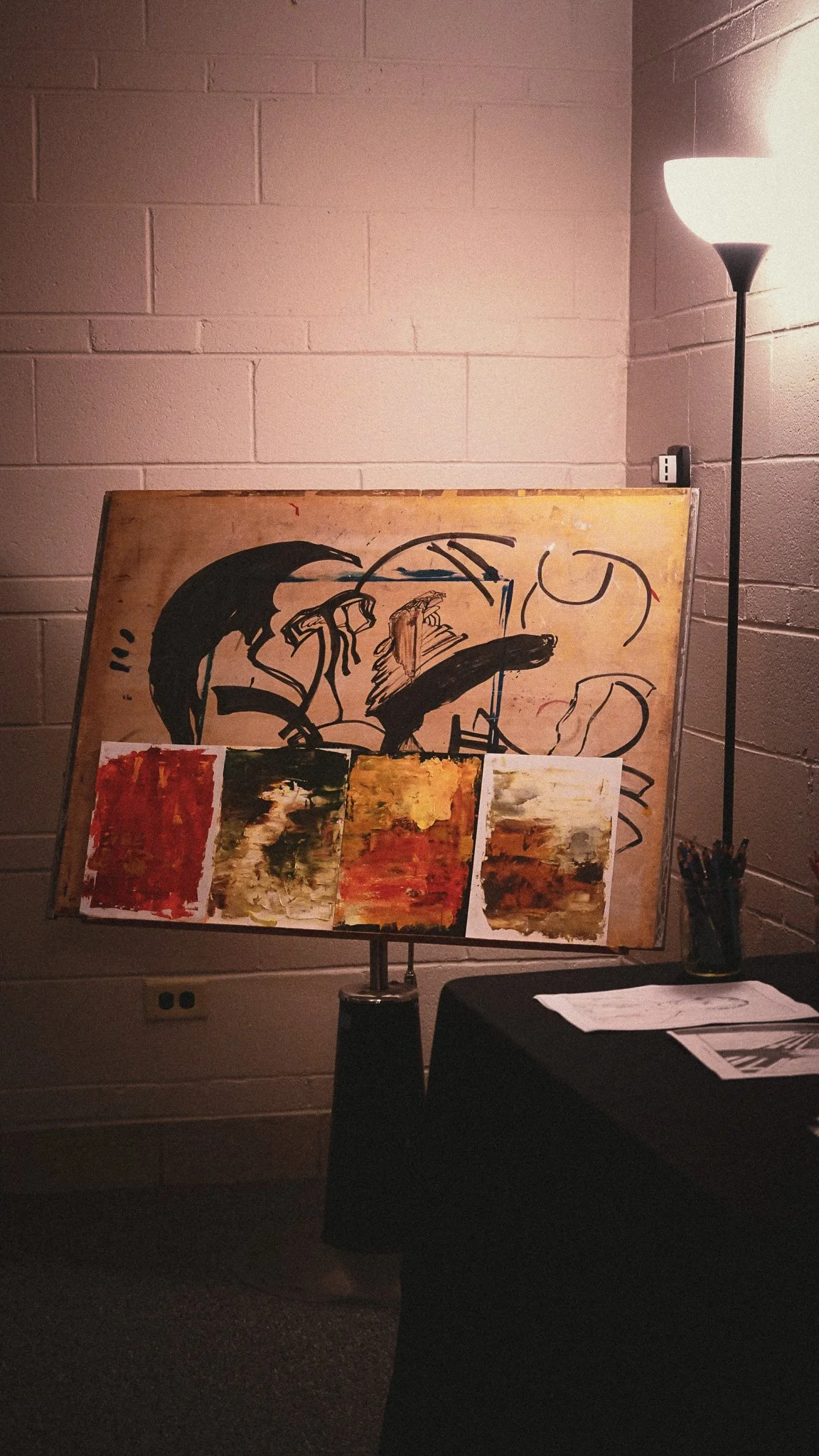 An art studio setup with an easel displaying both an abstract painting and a larger abstract on a wooden panel, illuminated by a tall floor lamp in a dimly lit room.