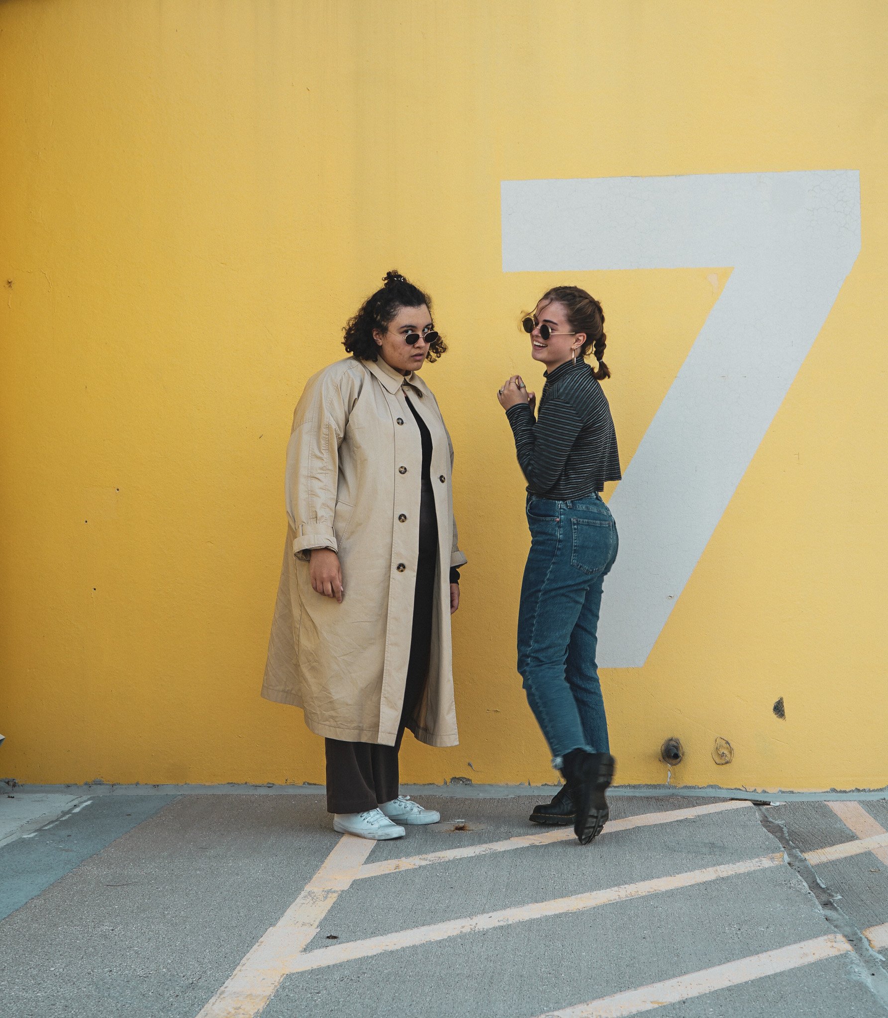 Two women wearing sunglasses, standing in front of a yellow wall with a large white number 7 painted on it, one smiling and the other looking serious.