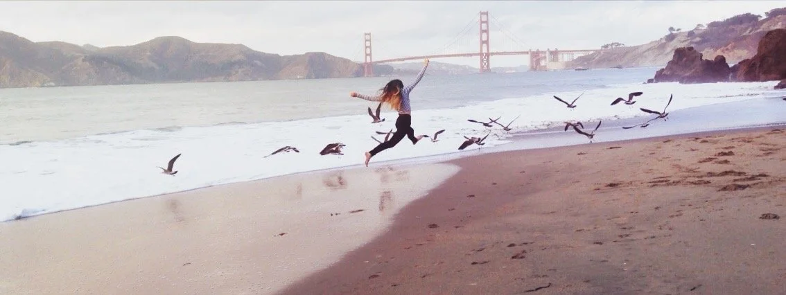 A woman jumping on a sandy beach near the ocean with seagulls flying around her. The Golden Gate Bridge is visible in the background.
