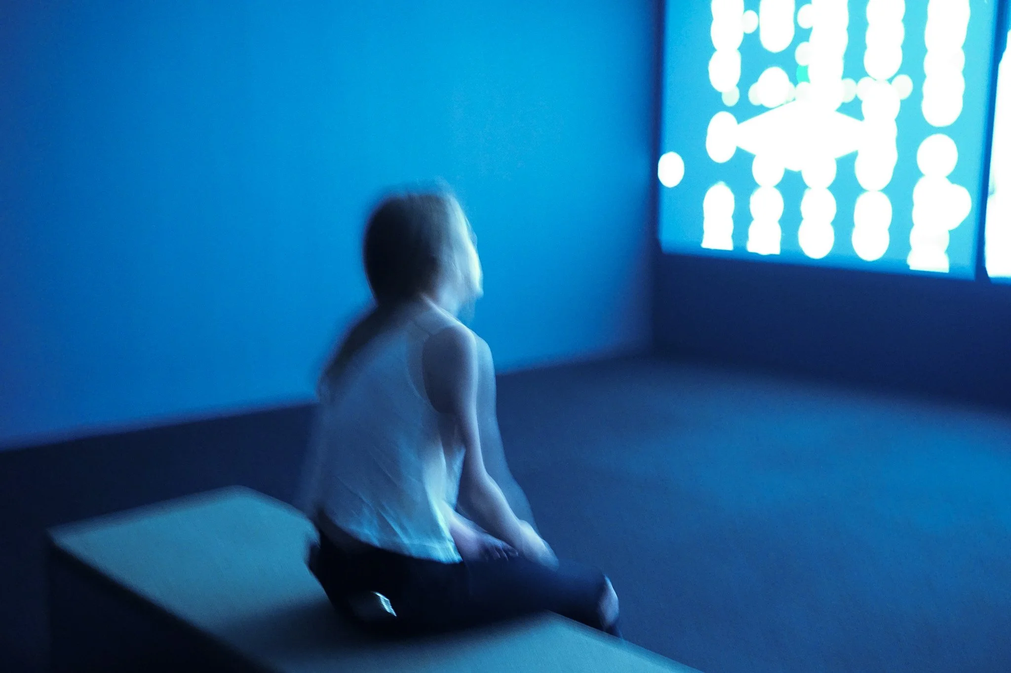 A girl sitting on a bench in a dark room, watching a large digital screen showing icons of people connected by lines, with a blue ambient light.