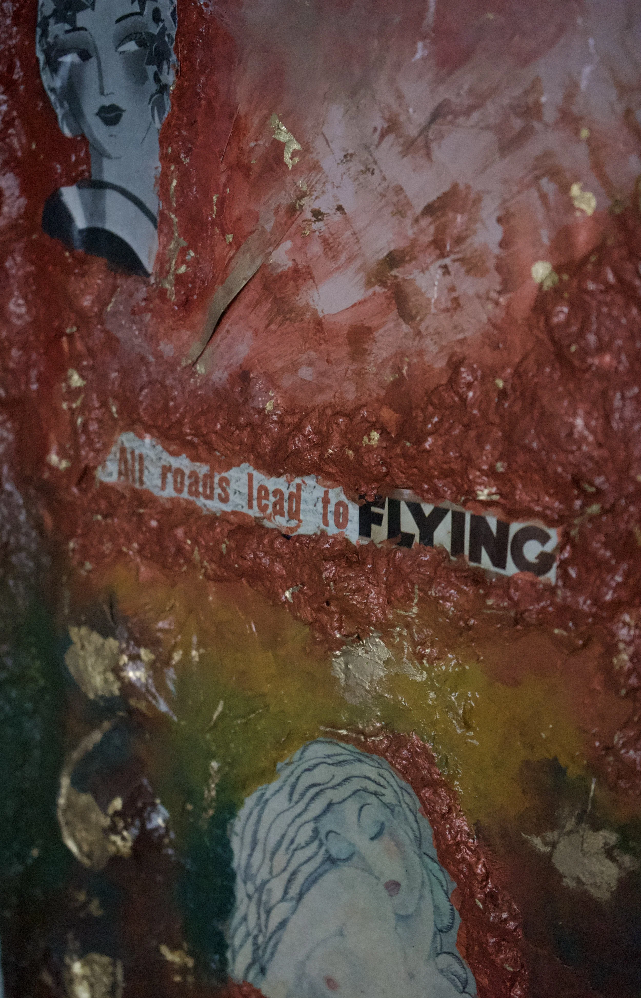 Close-up of a textured art piece with a collage of magazine cutouts, including a partial drawing of a woman's face and shoulder, with gold and red paint, and a torn piece of paper that reads 'All roads lead to FLYING'.