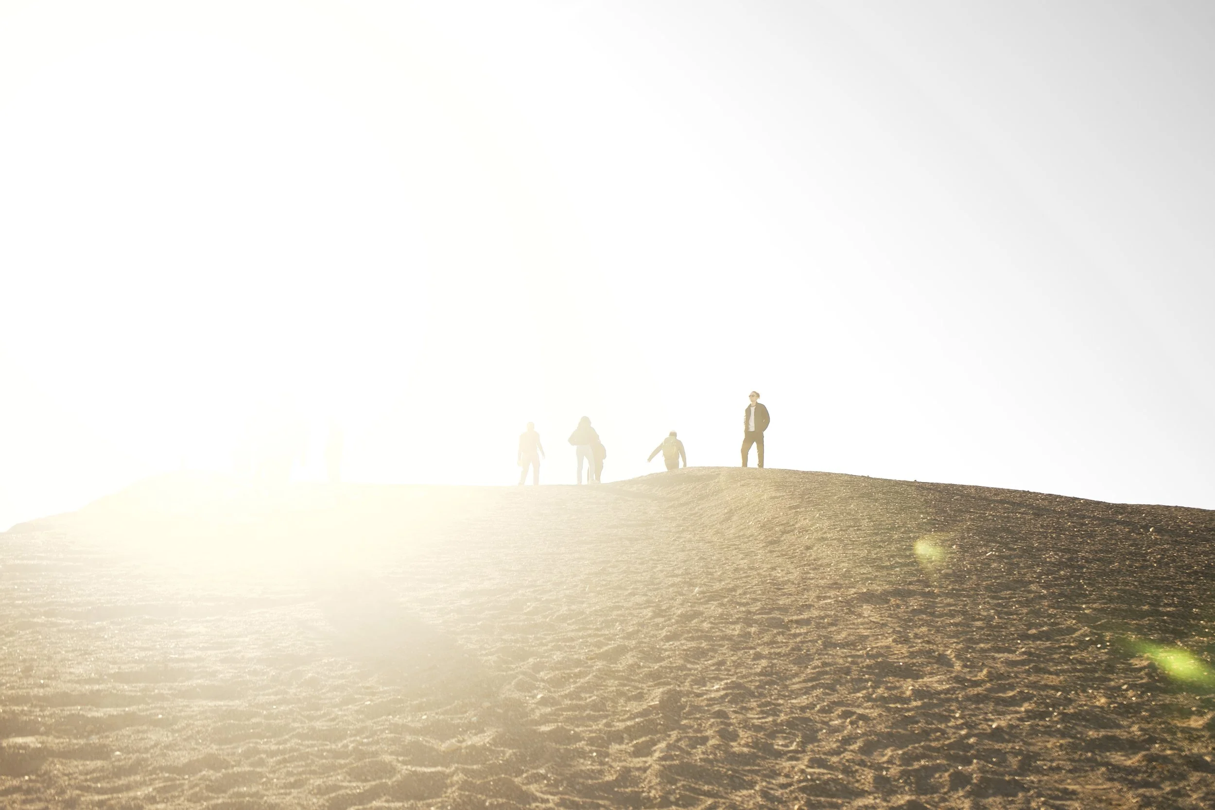 Group of people walking on a sandy hill at sunset or sunrise, with the sun bright and overexposed in the background.