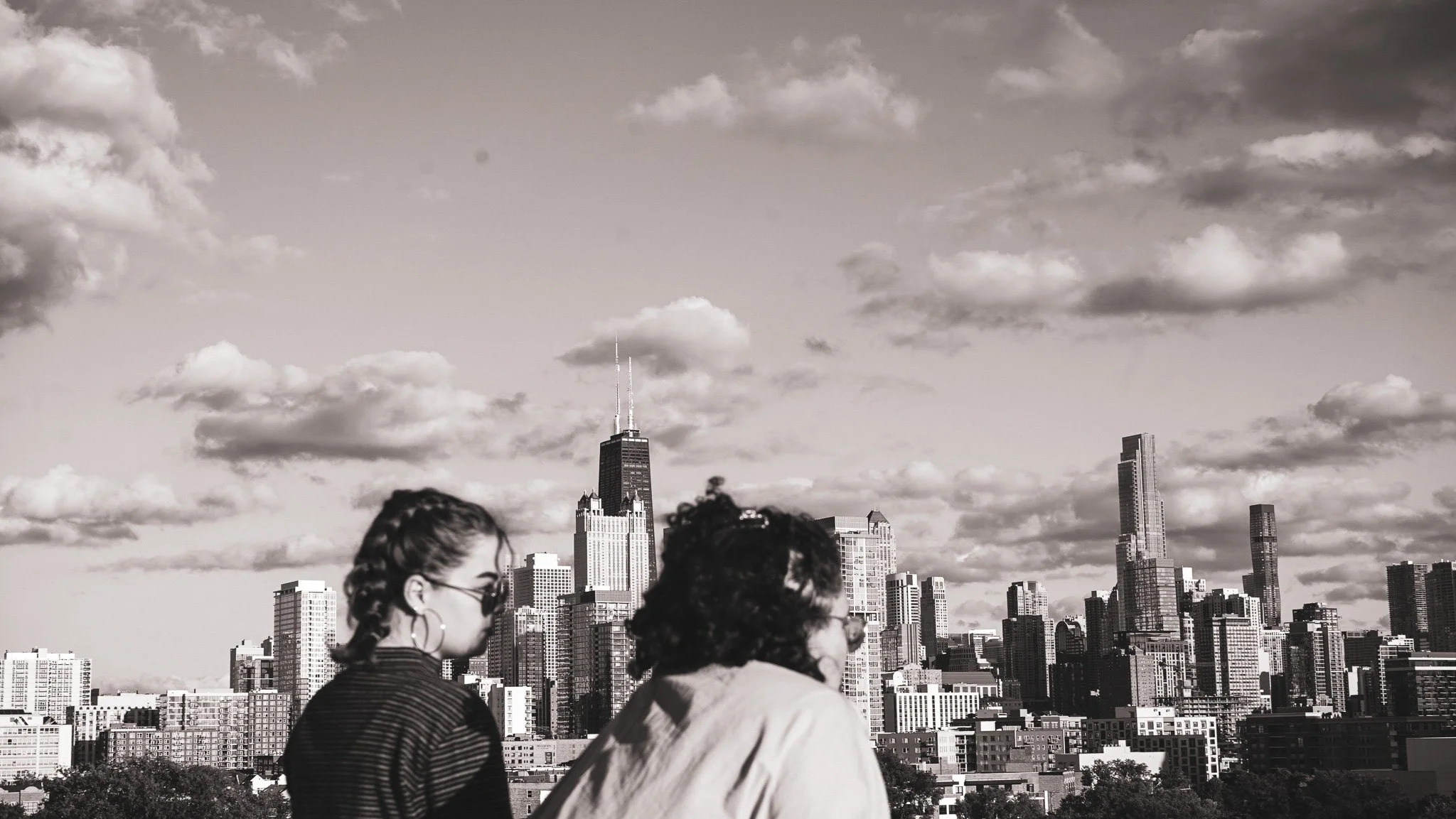 Two women with sunglasses in front of a city skyline with tall skyscrapers in black and white.