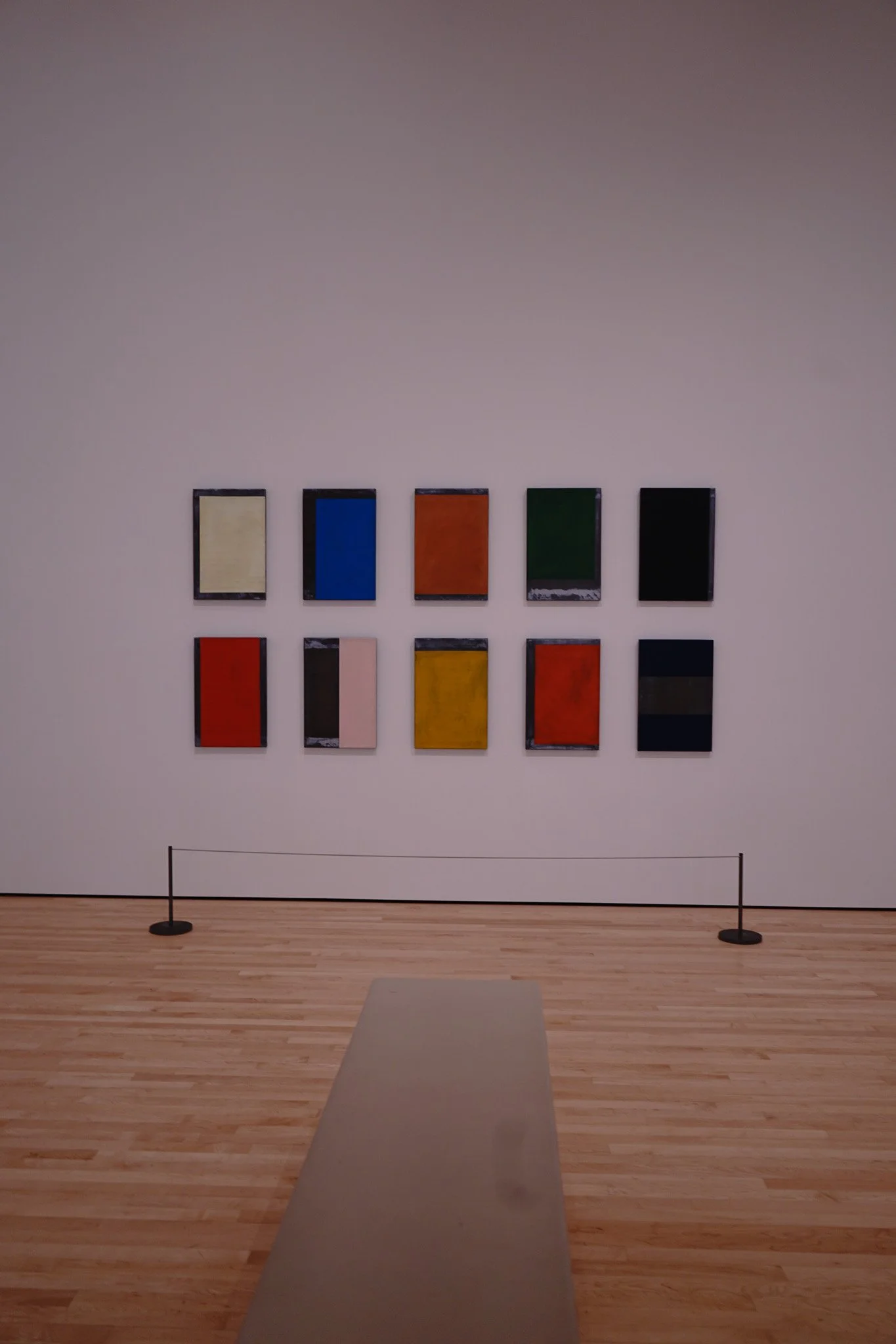 A museum gallery with ten abstract paintings on the wall arranged in two rows of five, and a beige bench in the foreground.
