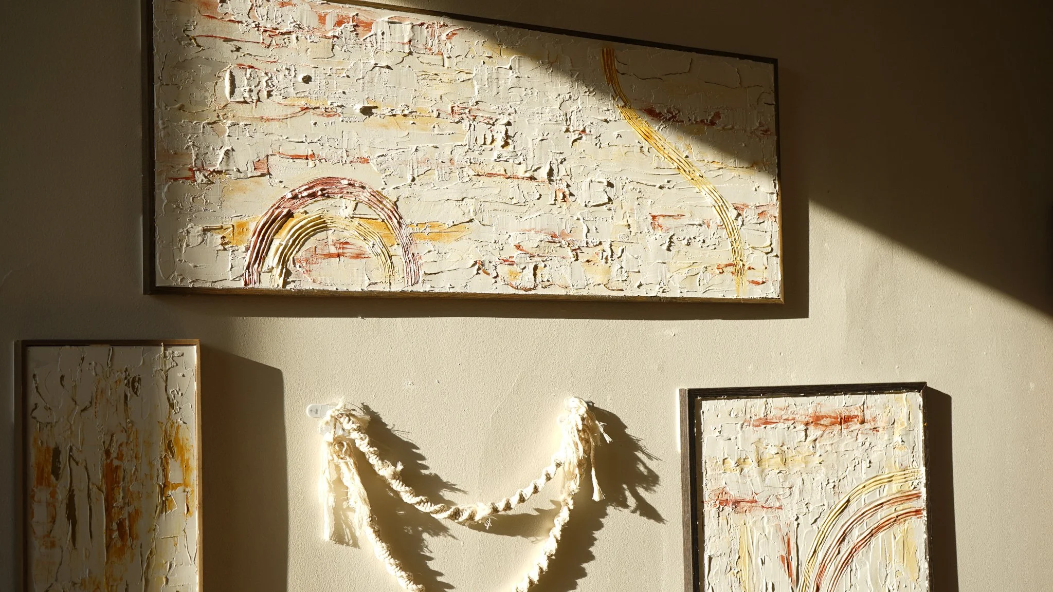 Decorative wall art with textured abstract paintings and a white rope hanging on a beige wall, illuminated by sun rays.