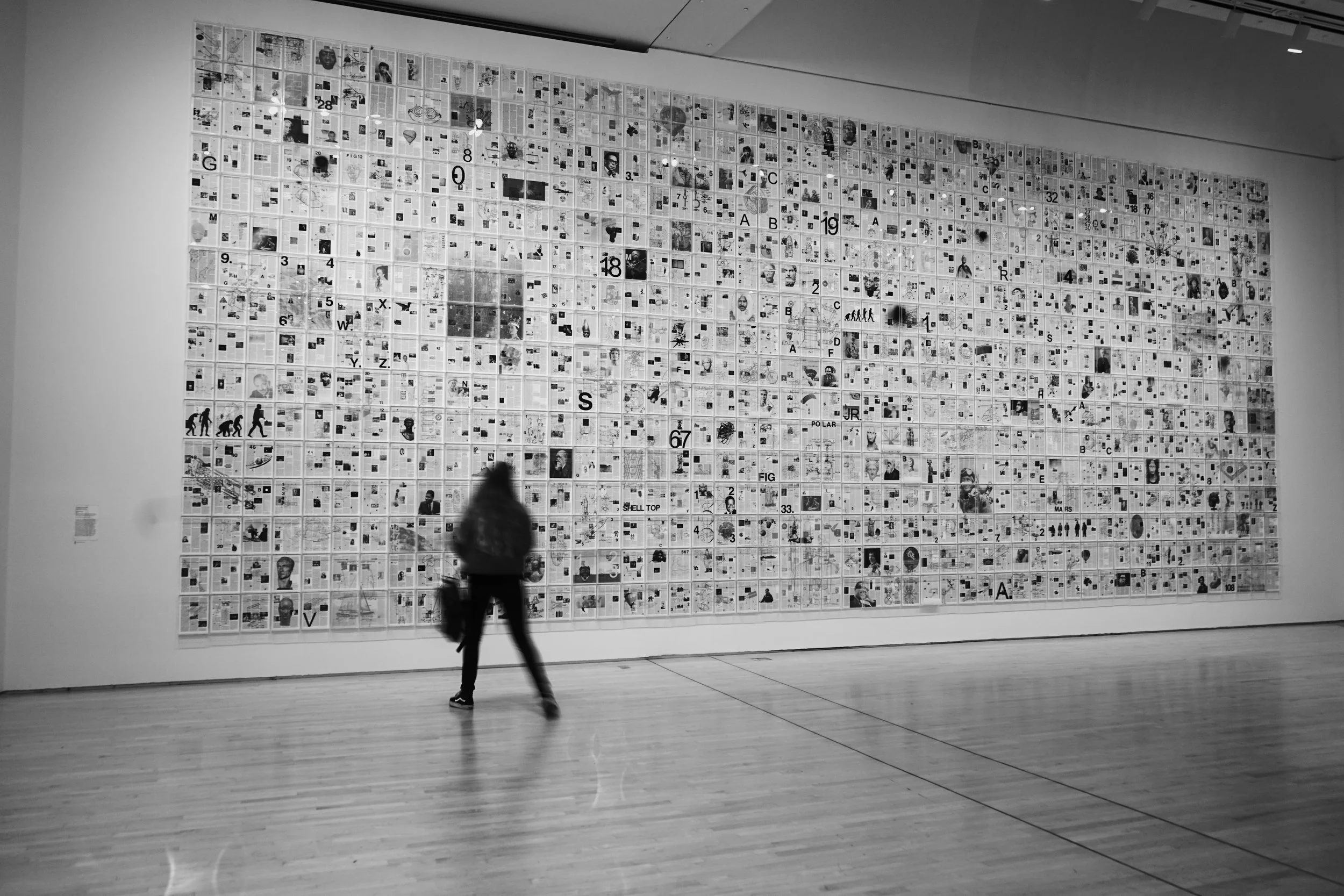 A person walking in front of a large collage of numerous small black-and-white images and text on a gallery wall.