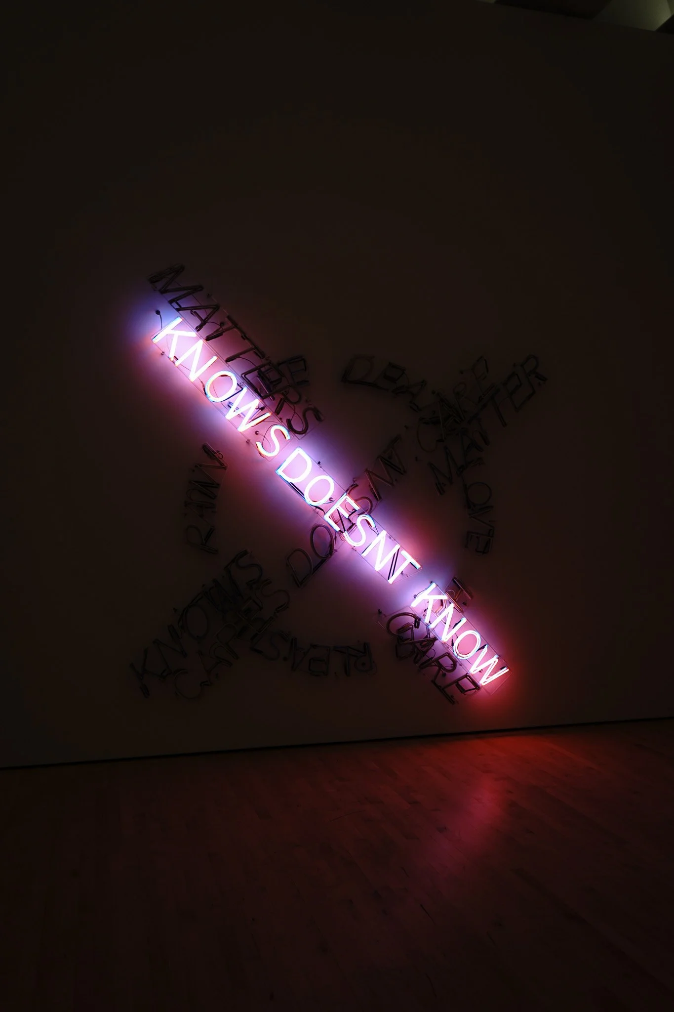 Pink neon sign with the words "KNOW'S DOESN'T KNOW" on a dark wall, reflected faintly on the wooden floor.