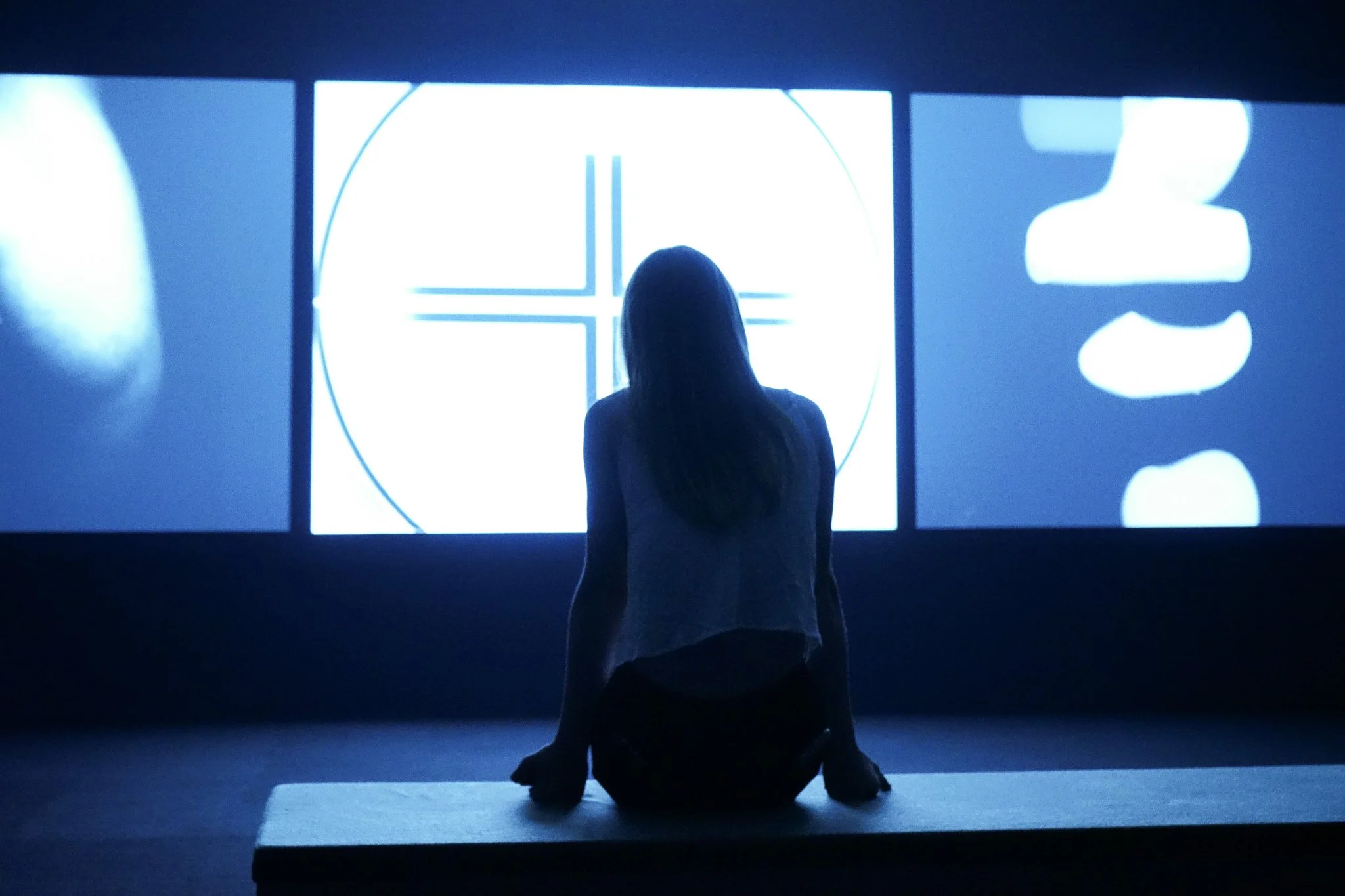 A woman sitting on a bench in front of three digital displays showing various abstract images and a target-like pattern, with the woman facing away from the camera in a dark room.