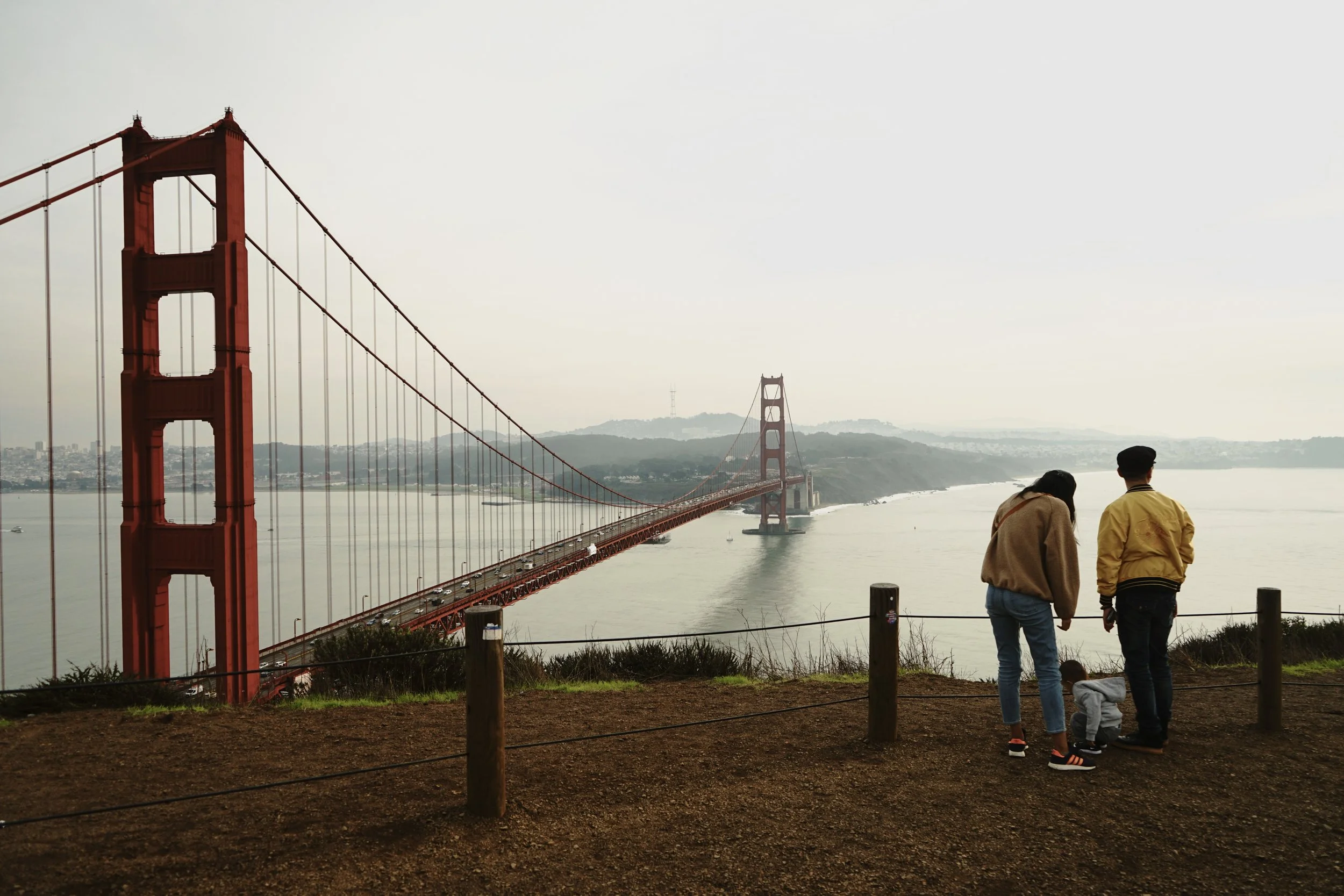 Three people, a woman, a man, and a child, standing near a fence on a hill overlooking the Golden Gate Bridge in San Francisco.