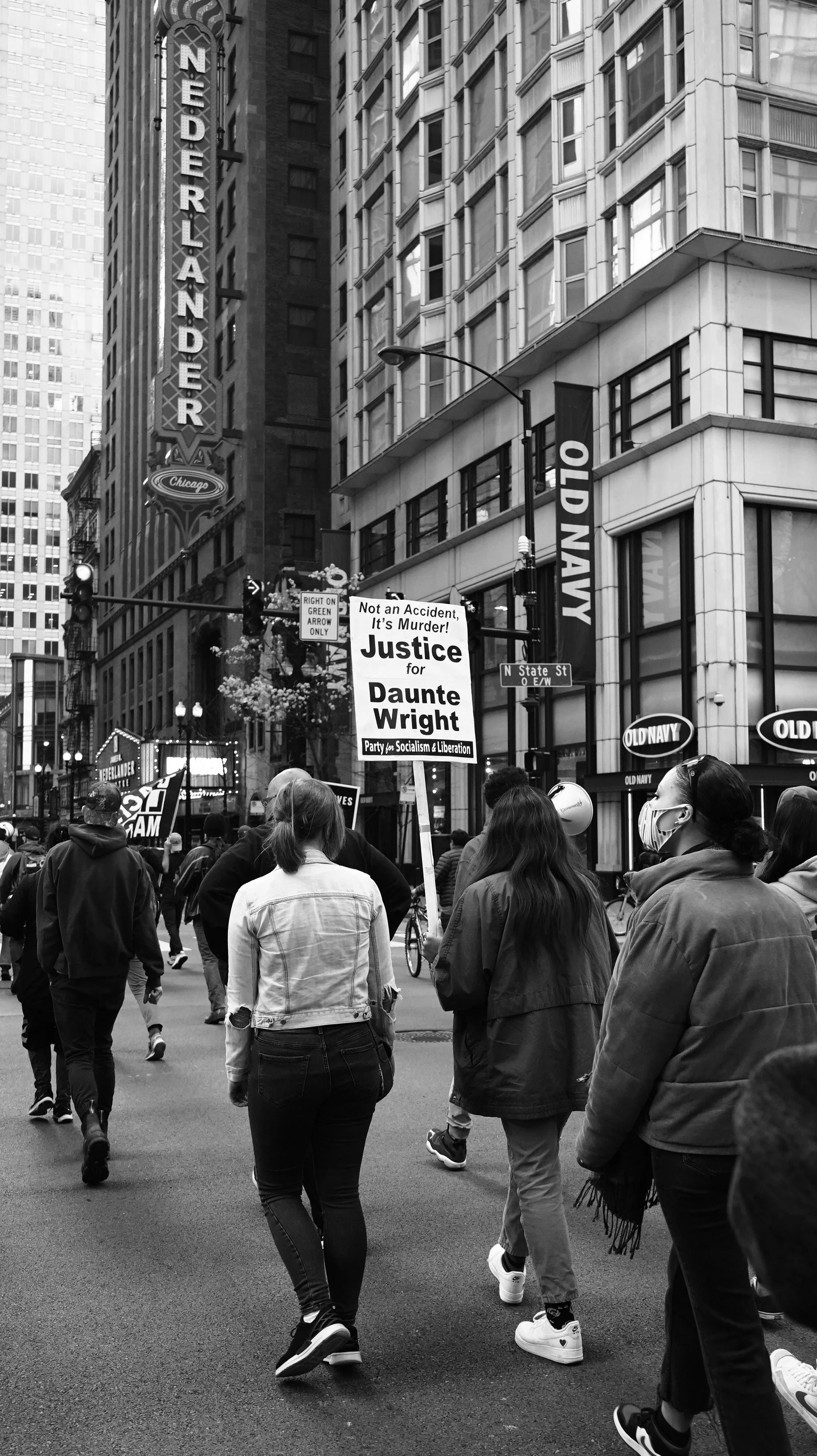 Protest in a city street with people carrying signs including one that says Justice for Daunte Wright, with tall buildings, a 'Nederlander' sign, and people wearing masks.