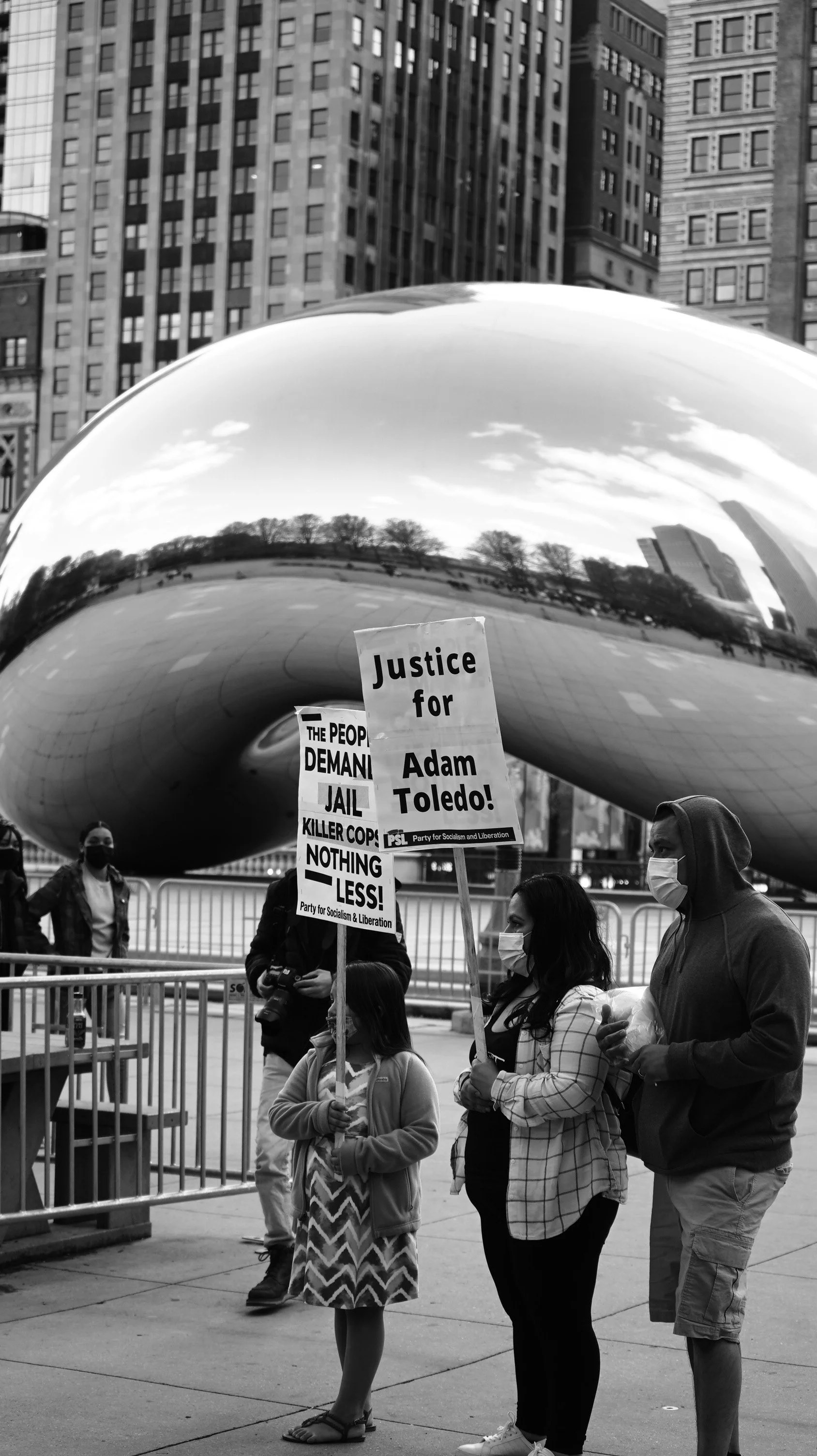 A group of people, including children, holding protest signs in front of Cloud Gate sculpture in Millennium Park, Chicago. The signs read 'Justice for Adam Toledo!' and 'The people demand jail, killer cops, nothing less!'. The scene is in black and w