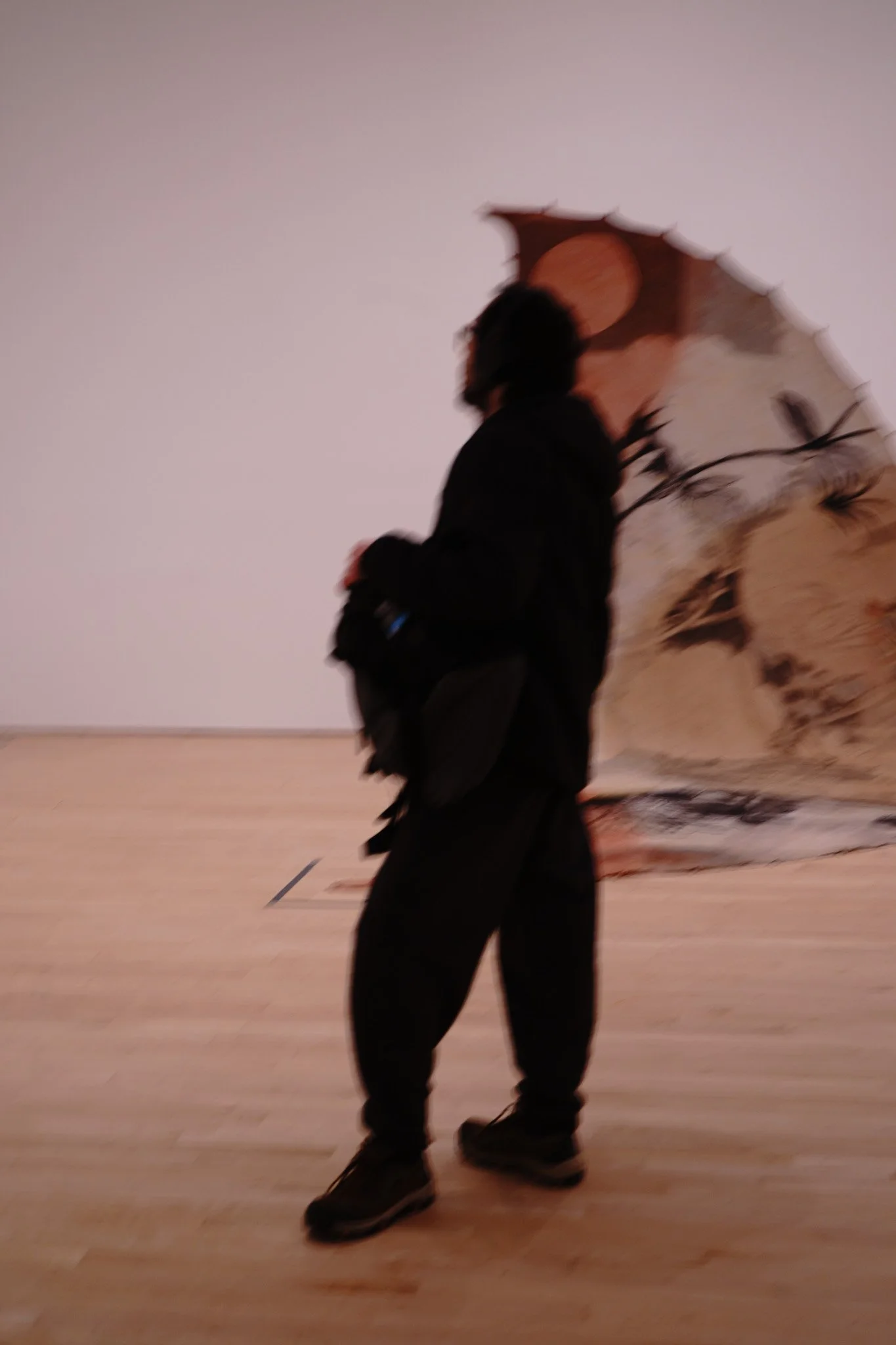 A person standing in front of a large abstract painting in an art gallery. The person is dressed in dark clothing and holding an umbrella, with their profile visible against a pale wall and wooden floor.