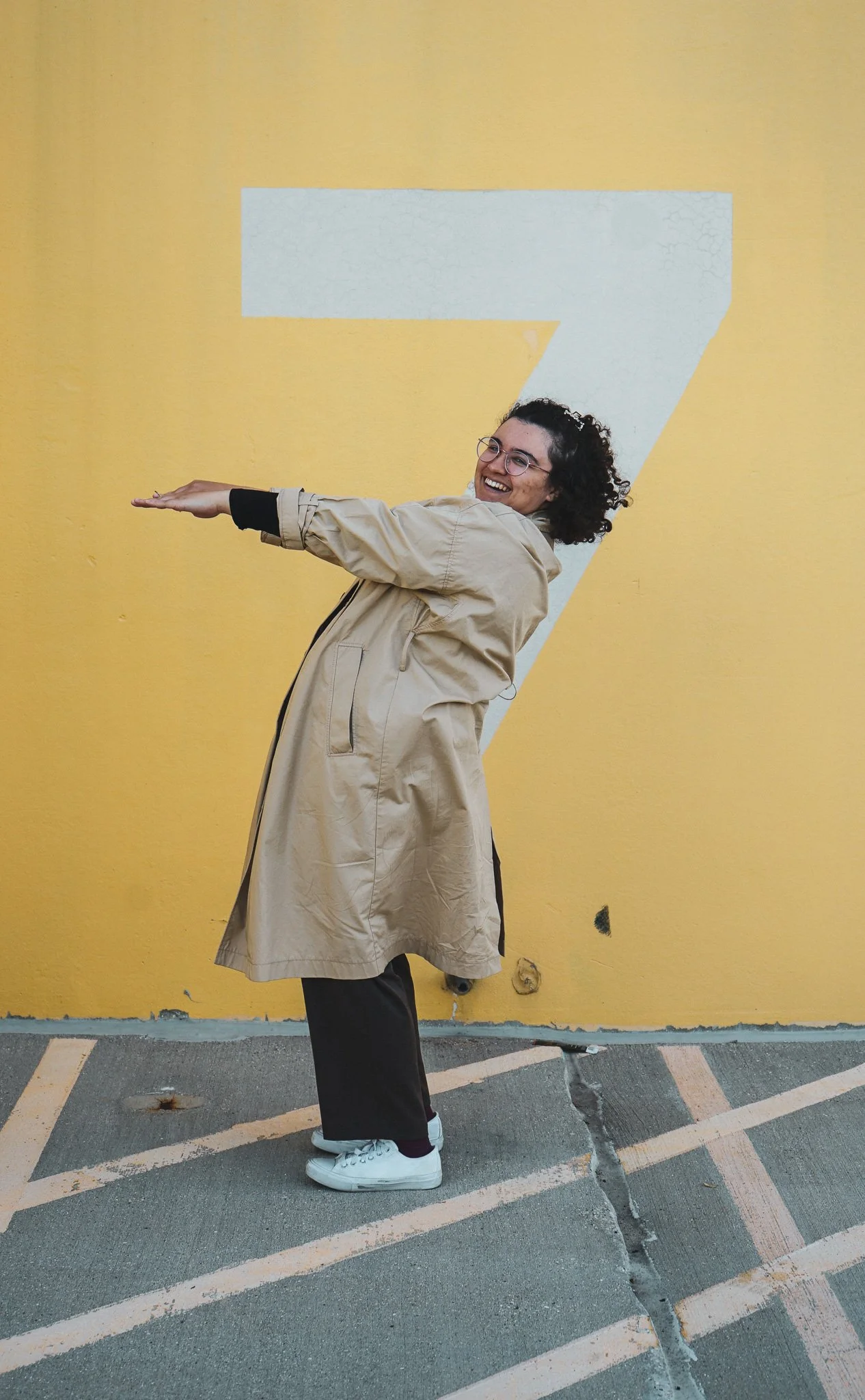 A woman in a beige trench coat, black pants, white sneakers, and glasses, smiling and pointing forward, standing in front of a yellow wall with a large white number seven.