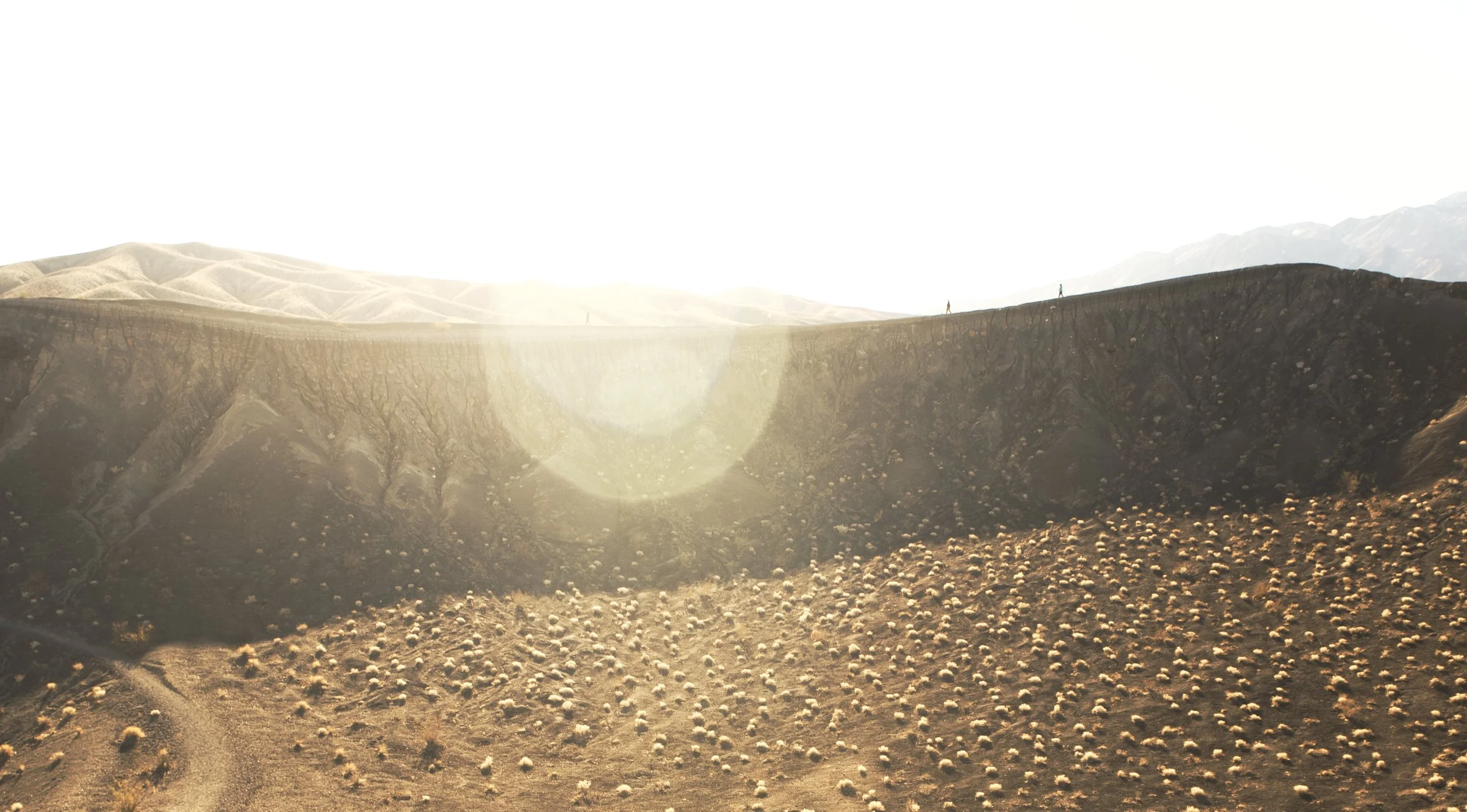 A barren, desert landscape with sparse vegetation and rolling hills in the background, illuminated by bright sunlight.