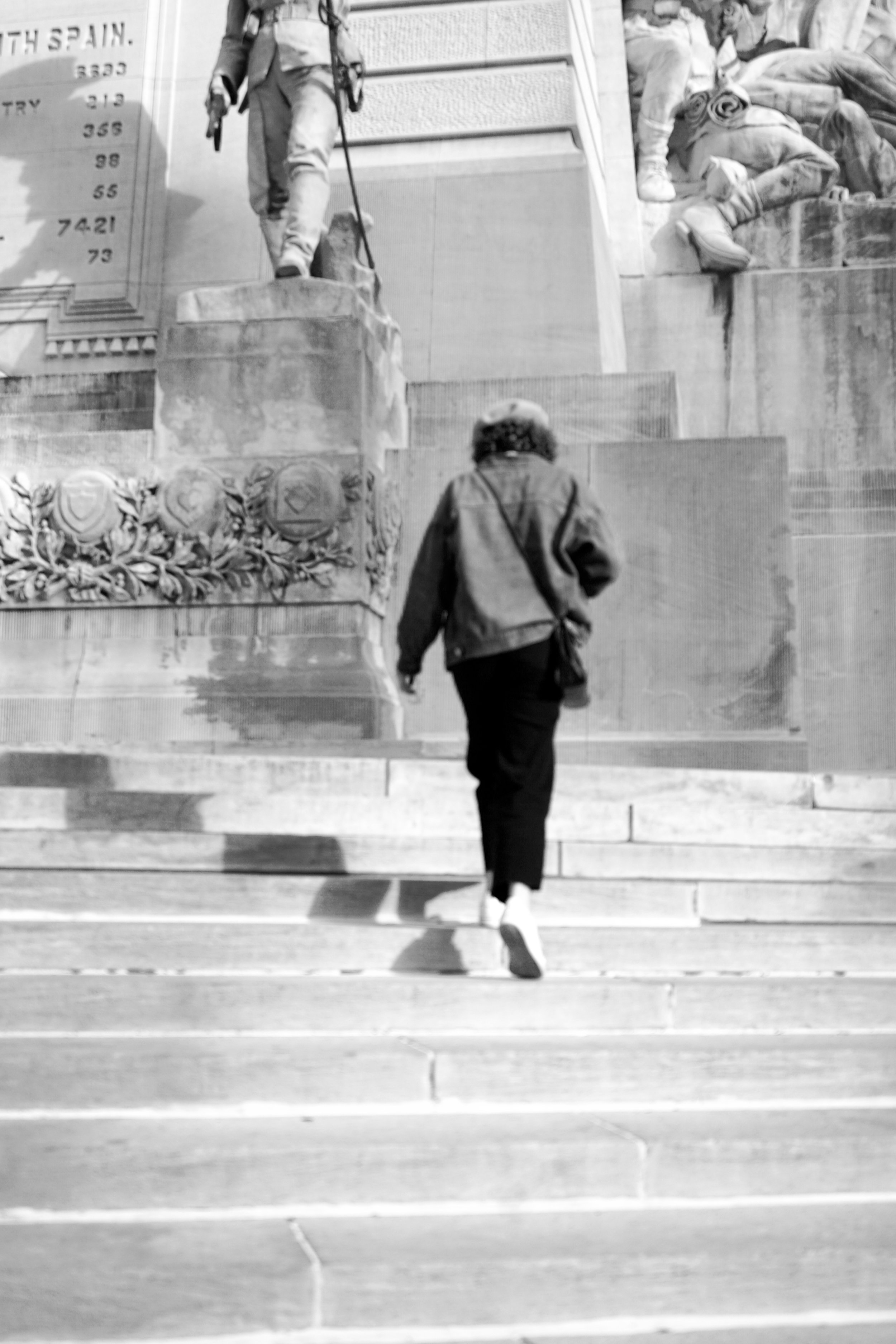 A woman walking up steps toward a monument or statue with detailed carvings and sculptures, including figures and decorative elements, in black and white.
