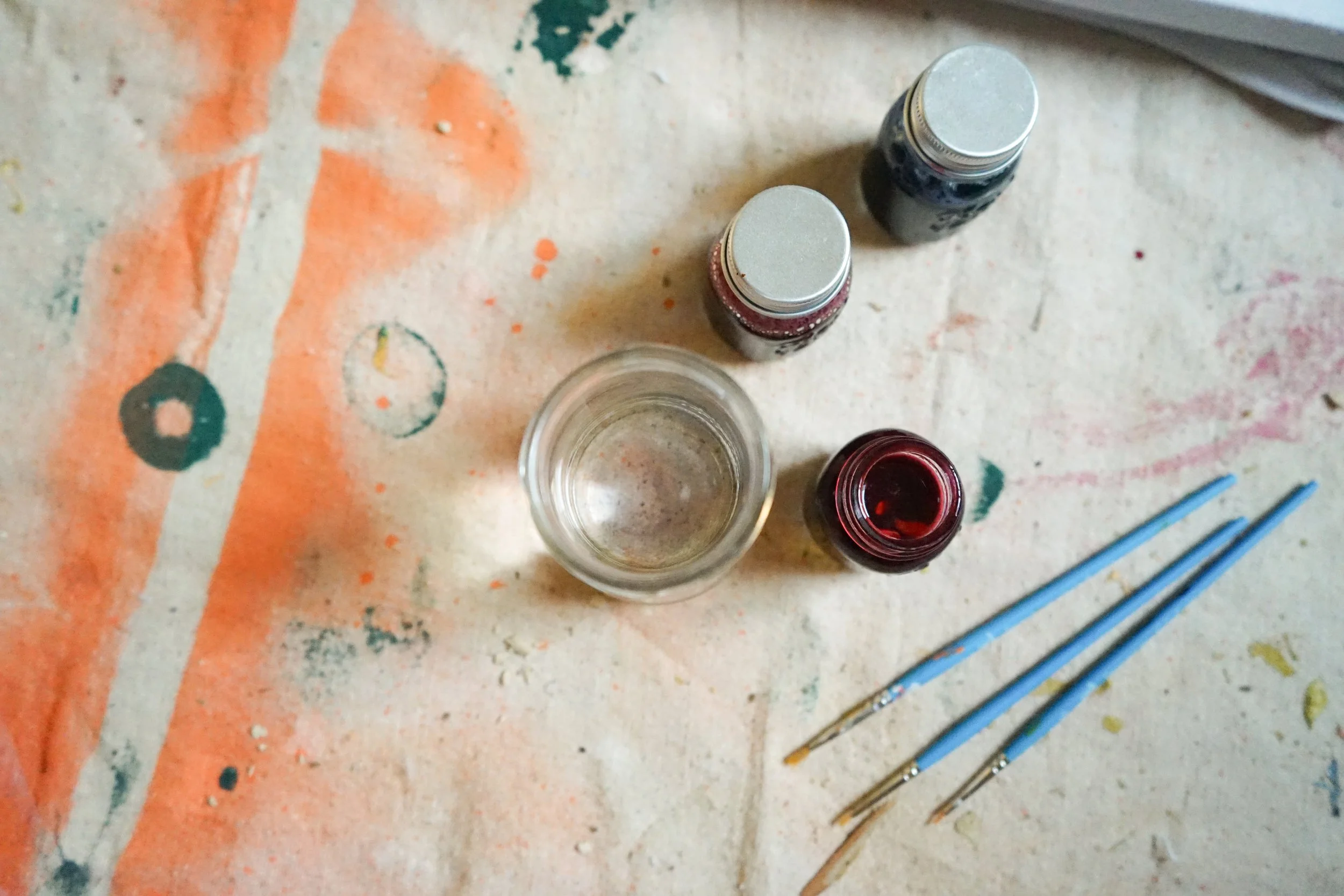 Paint bottles, a glass of water, and paintbrushes on a colorful, stained tabletop.