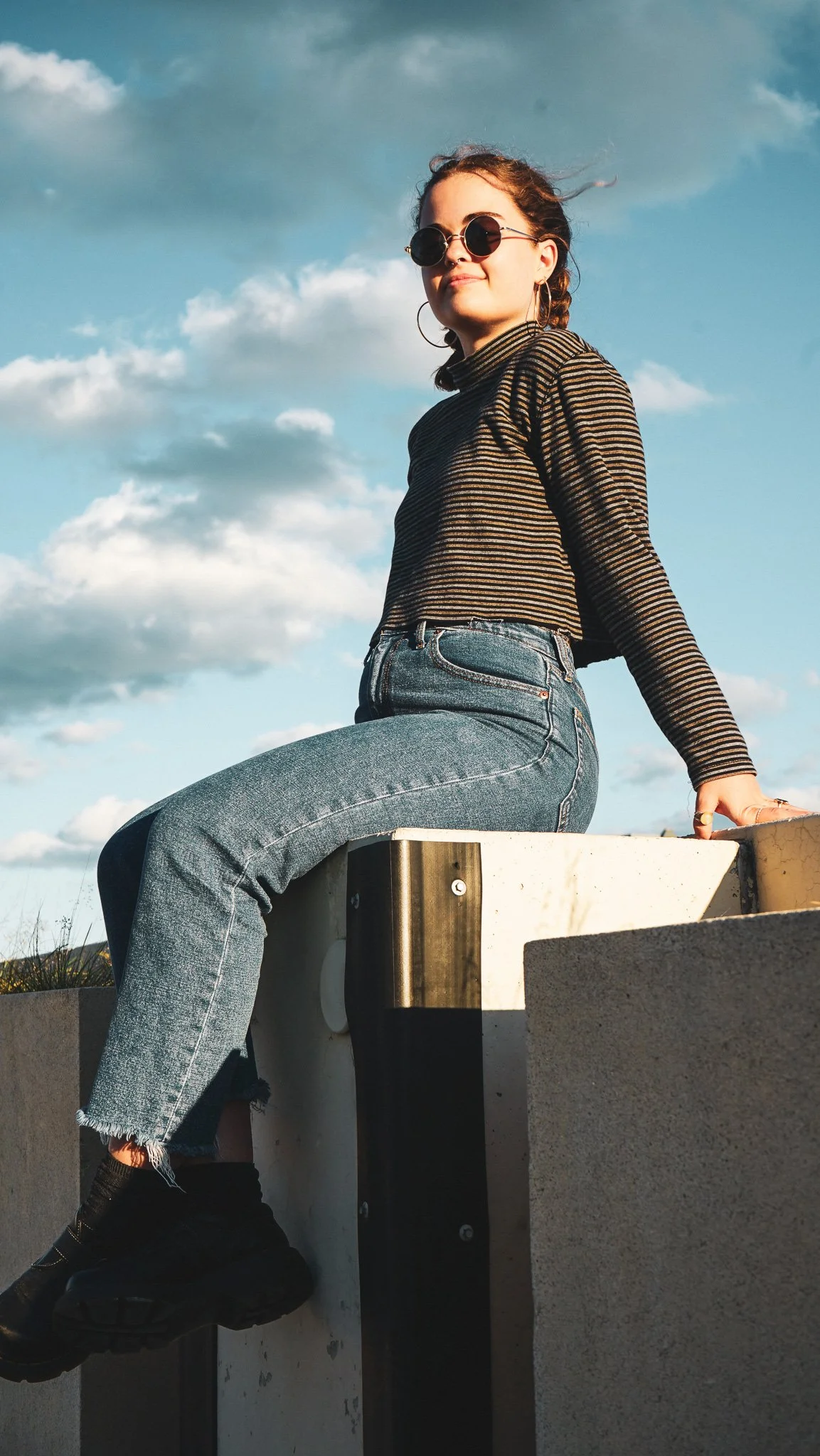 A young woman with curly hair and sunglasses sitting on a concrete block outdoors with a partly cloudy sky in the background.