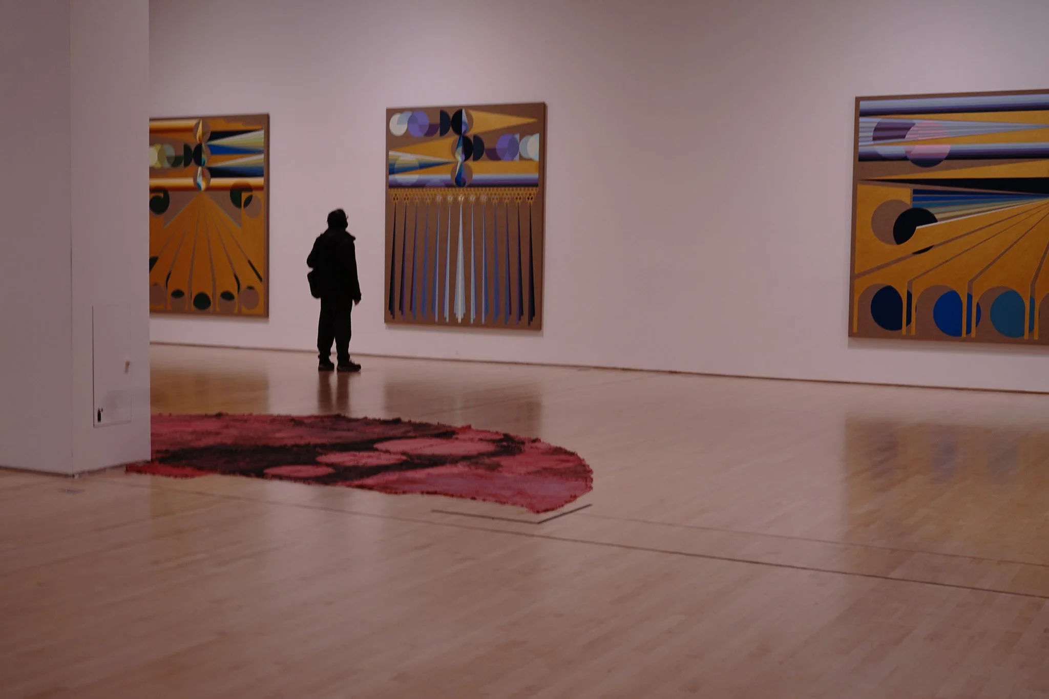 A person standing in an art gallery, viewing abstract paintings on the wall, with a red and black rug on a wooden floor.