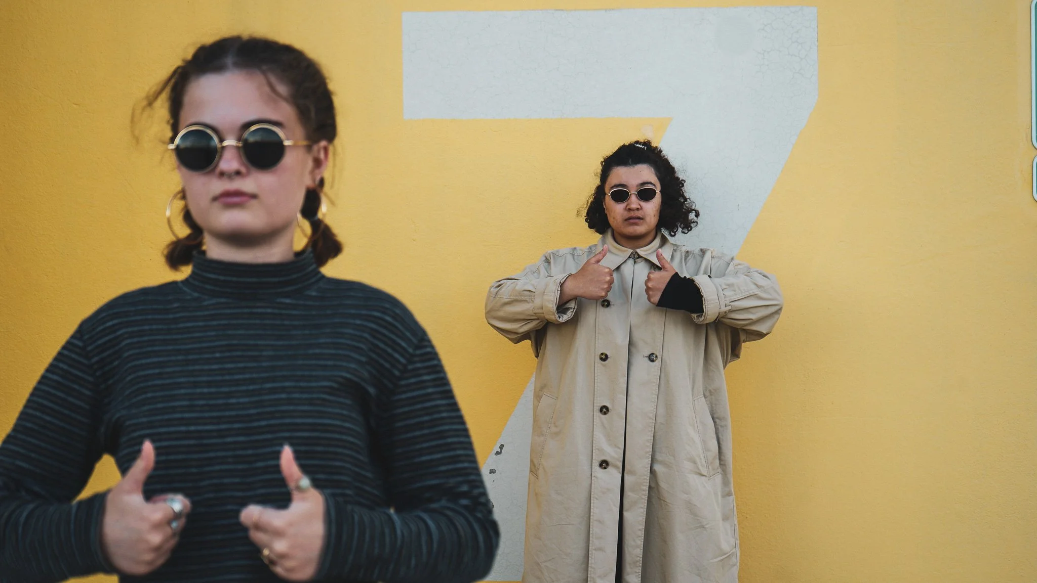 Two women wearing sunglasses and casual clothing stand against a yellow wall with a gray and white geometric design. The woman in the foreground has short hair, round sunglasses, and a striped turtleneck, with her thumbs up. The woman in the backgrou