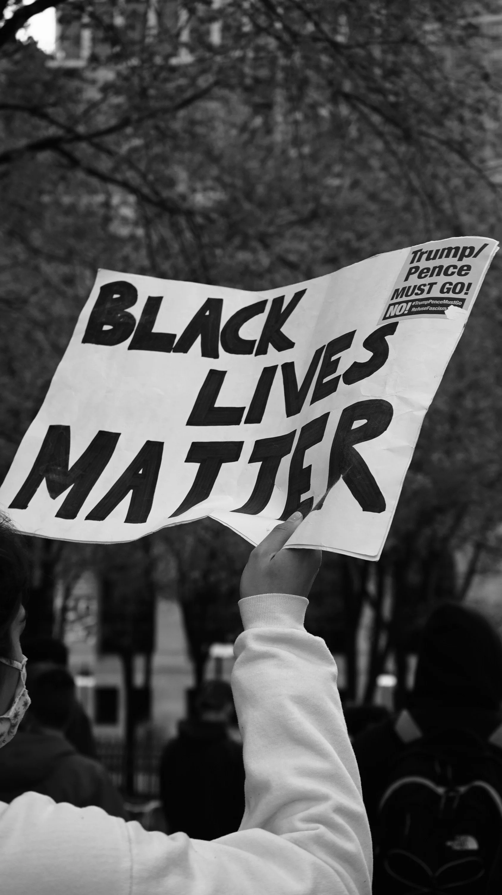 A person holding a sign that reads 'Black Lives Matter' during a protest, with other protesters and trees in the background.