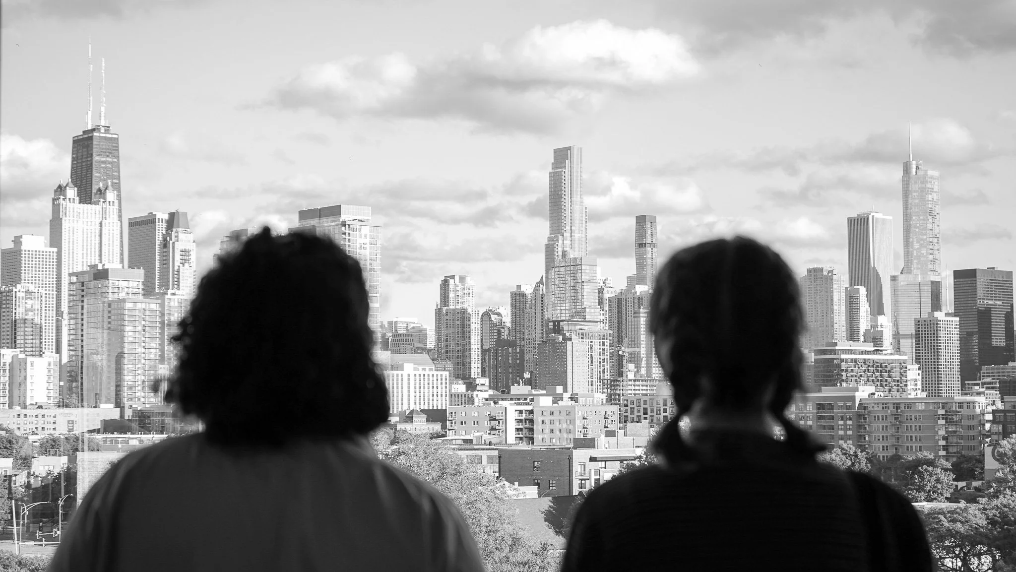 Two people sitting and looking at a city skyline with tall buildings and skyscrapers in the background, in black and white.